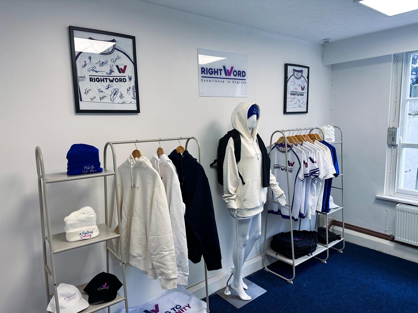 Display of white and blue clothing and accessories with RightWord branding in a store, including hoodies, t-shirts, hats, and a mannequin dressed in a hoodie with a backpack, all arranged on racks and shelves.
