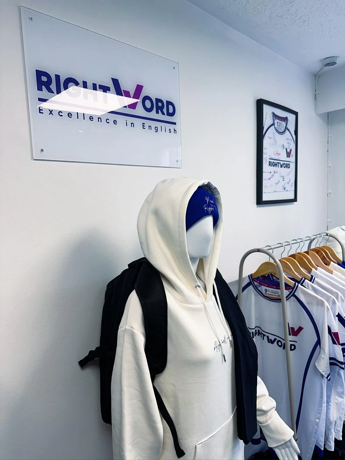 Display of sports jerseys and a mannequin with hoodie and backpack in a store or exhibition area, with a sign reading 'Right V Word' and slogan 'Excellence in English' on the wall.