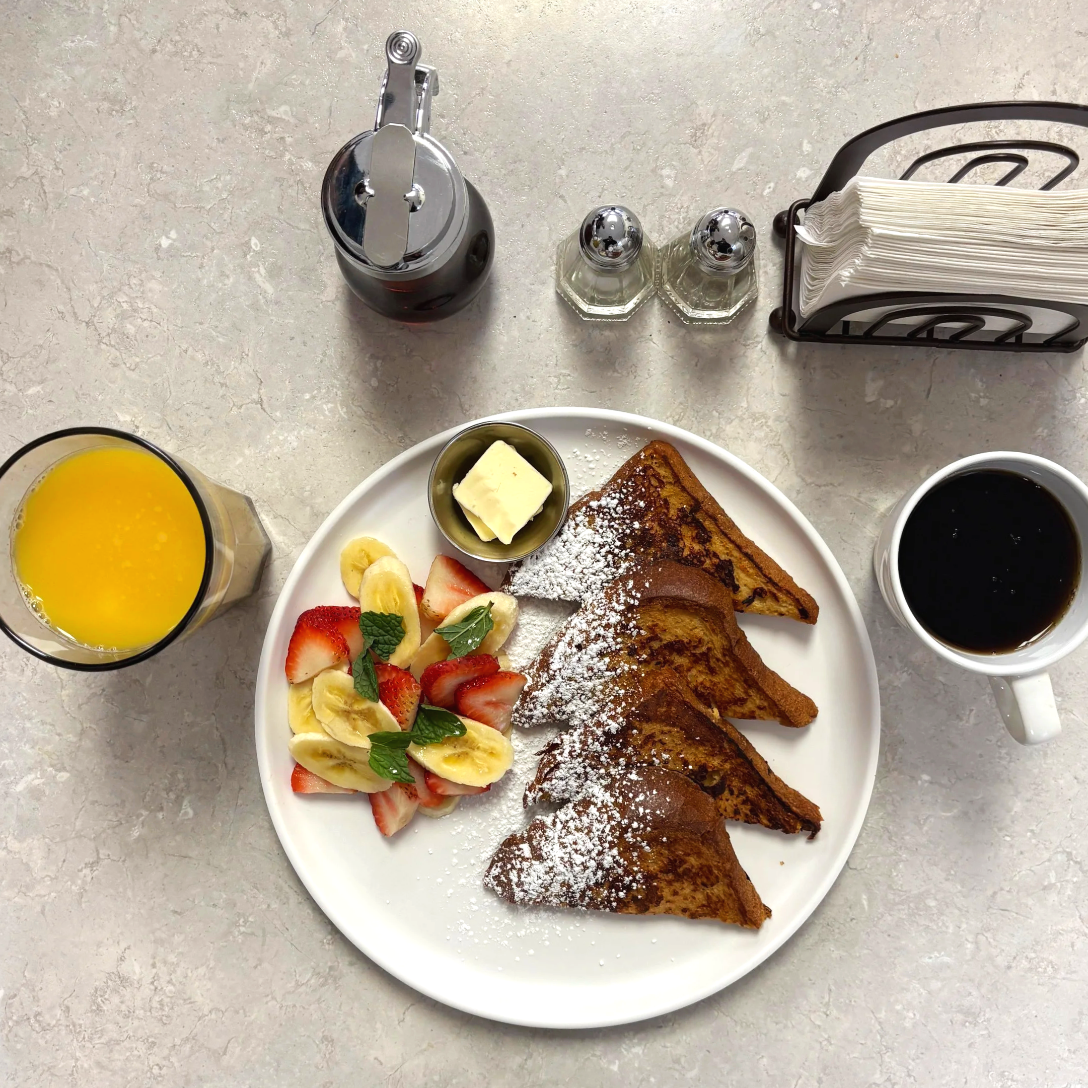 Plate of French toast garnished with powdered sugar, accompanied by a side of sliced strawberries and bananas, topped with mint leaves, with a small dish of butter, a glass of orange juice, and a cup of black coffee on a breakfast table.