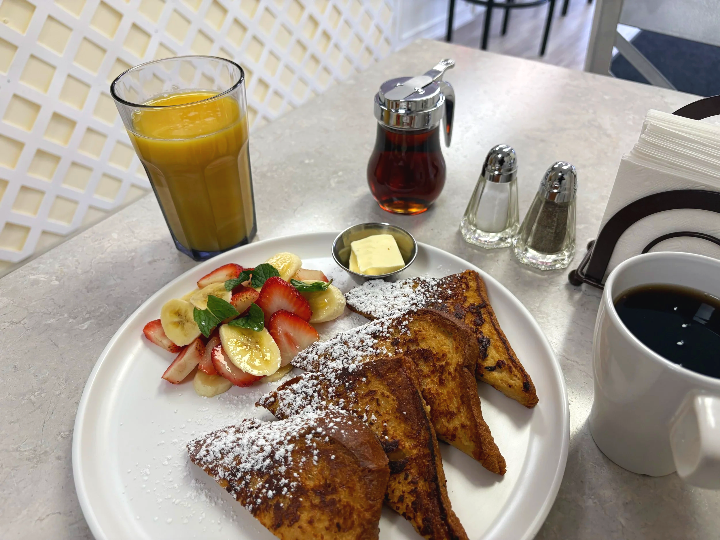 A breakfast plate with French toast, fresh fruit salad, a small dish of butter, and powdered sugar. Beverages include a glass of orange juice and a cup of black coffee. There are salt and pepper shakers, a small syrup dispenser, and napkins on the ta