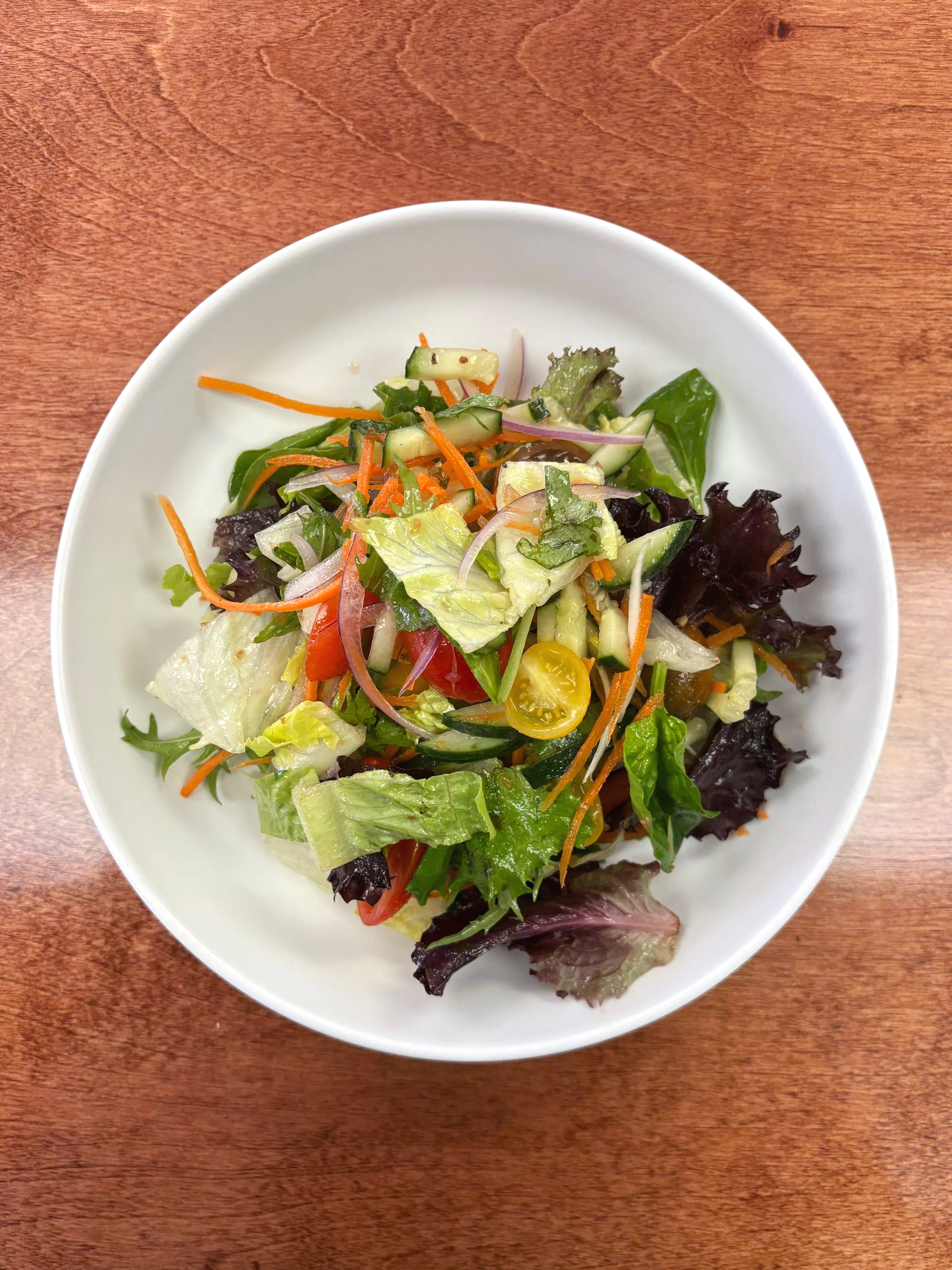 A white bowl filled with mixed green salad including lettuce, cherry tomatoes, cucumbers, shredded carrots, and red onions on a wooden table.