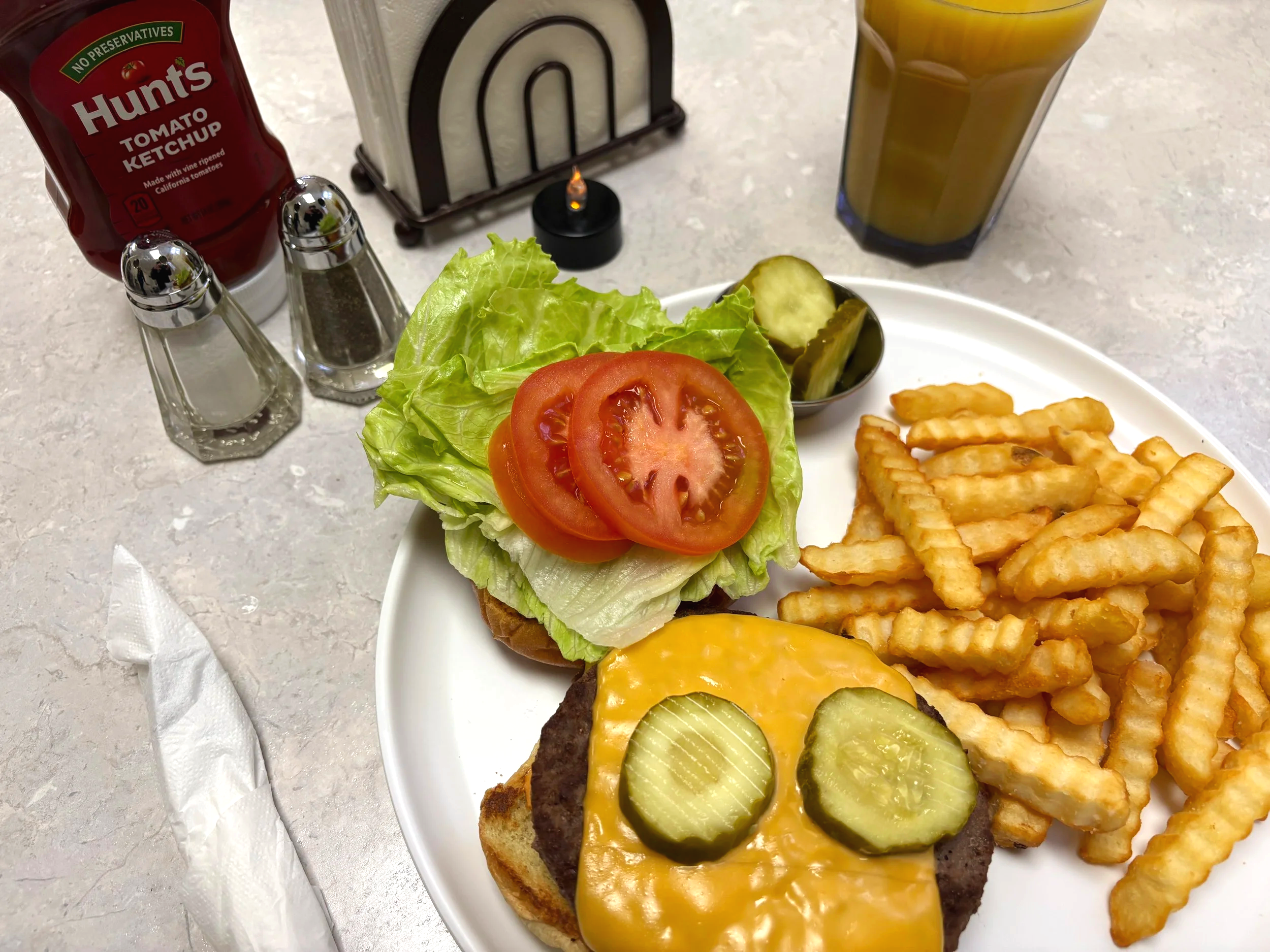 A plate of cheeseburger with pickles, lettuce, and tomato, crinkle-cut French fries, and a small side of pickles. In the background, there is a glass of iced tea, a bottle of Hunt's ketchup, and condiments shakers, with napkins and a toothpick holder