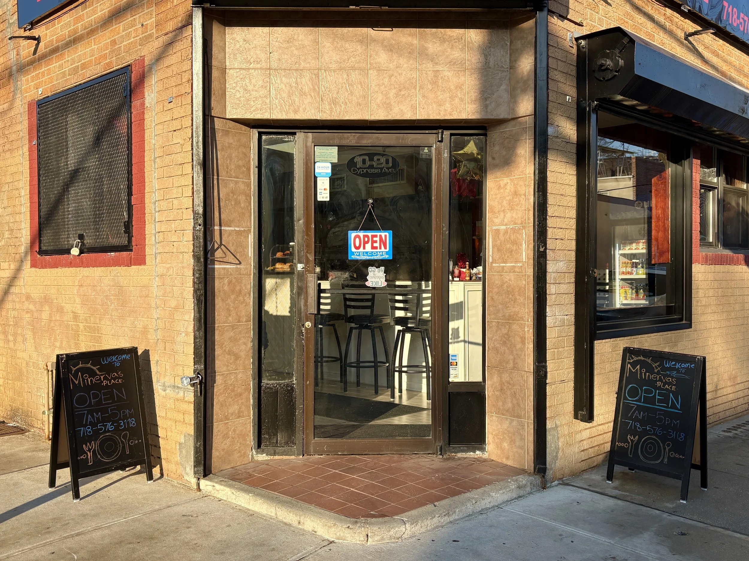 The storefront of a small business with a glass door that has an 'Open' sign hanging on it. There are two black chalkboard signs outside, one on each side of the entrance, displaying business hours and welcoming messages. The building has a brick exterior with black trims around the door and windows. The interior visible through the glass shows chairs and a counter.