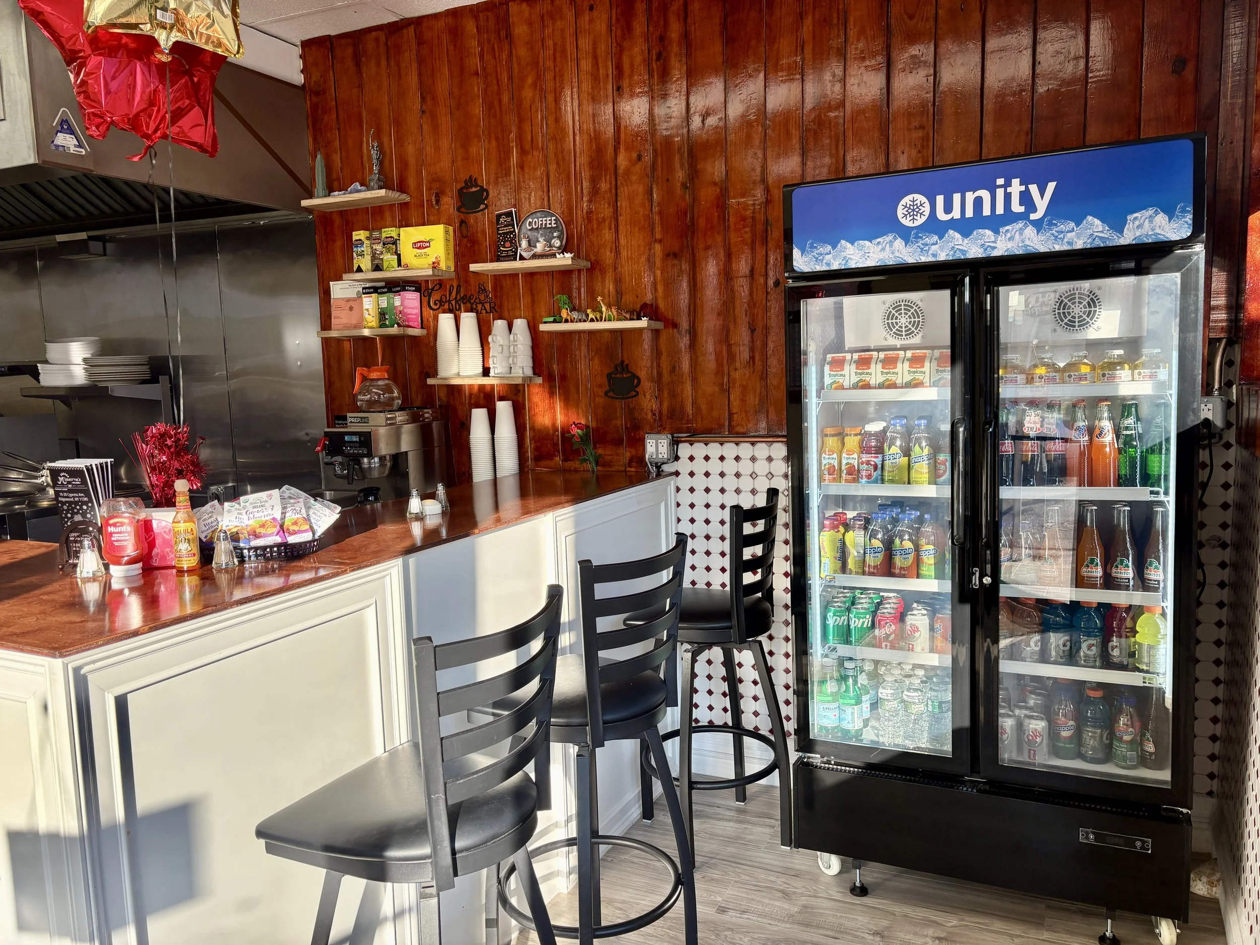 Interior of a cafe or small restaurant with a wooden wall, shelves holding coffee supplies, a glass refrigerator stocked with beverages, and seating with black chairs.