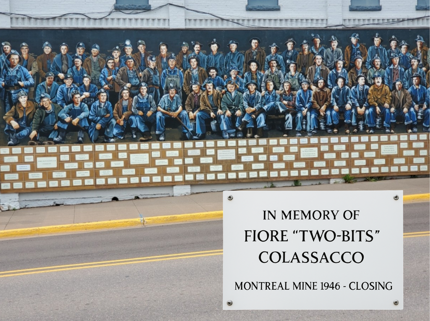 A mural featuring numerous miners in blue and brown work clothes, hats, and glasses, with some holding tools, situated above a row of plaques on a brick wall. A white sign in front of the mural reads, "In memory of Fiore 'Two-Bits' Colasacco, Montreal Mine 1946 - Closing."