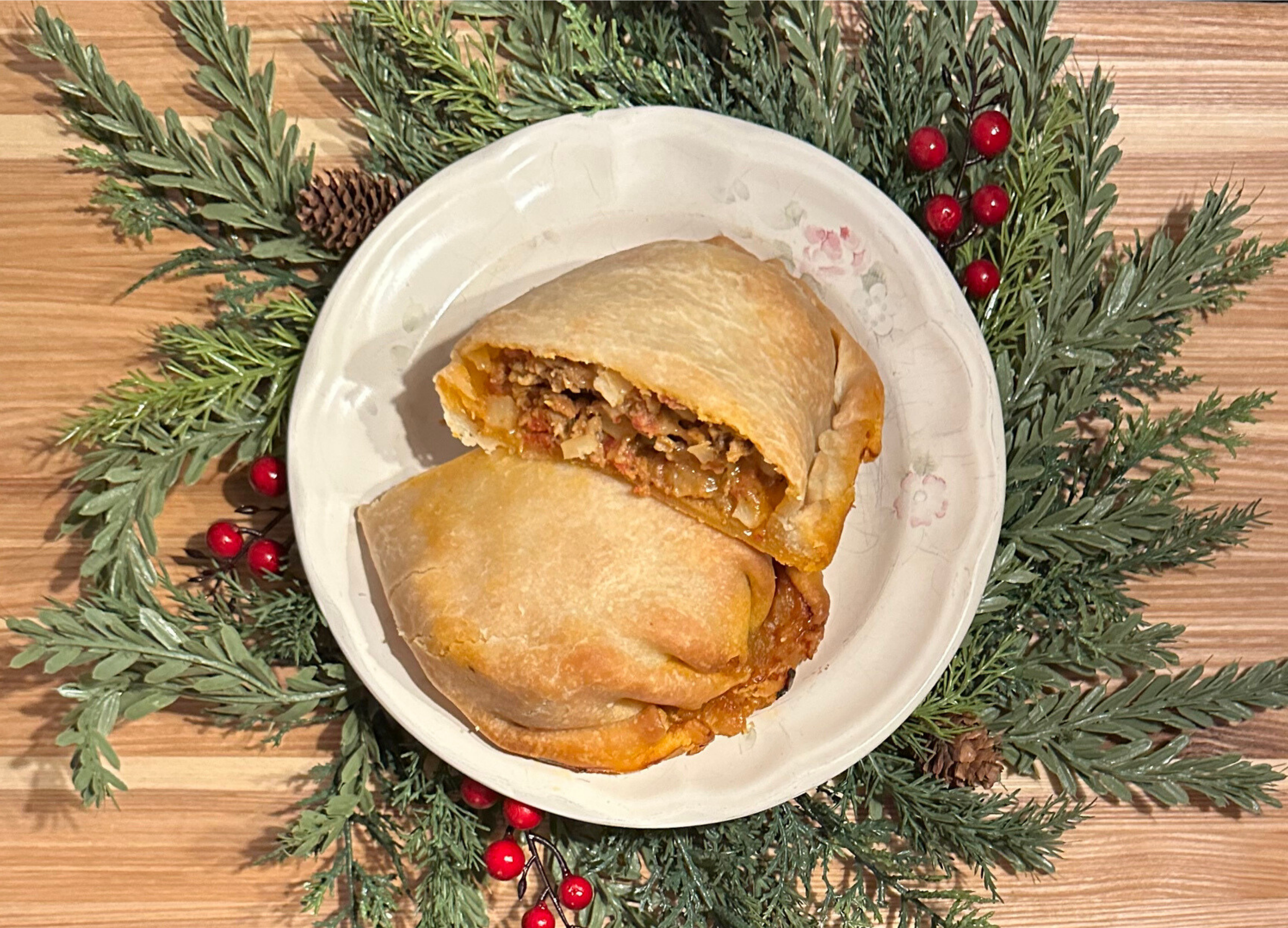 A plate with two egg rolls, one sliced open to show beef, vegetables, and sauce inside. The plate is surrounded by green pine branches and red berries, on a wooden surface.