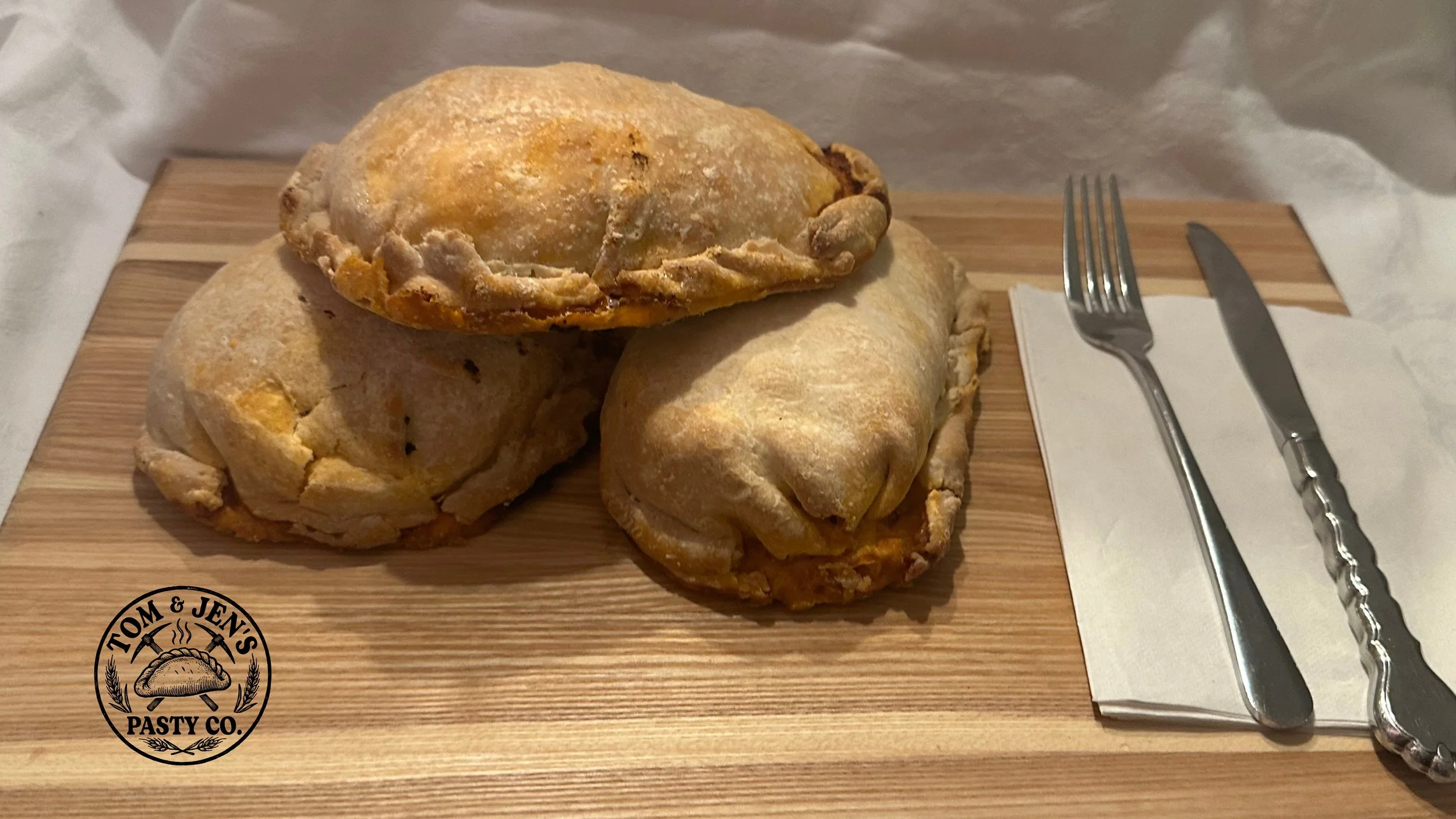 Three baked pastry empanadas on a wooden serving board with a fork and knife on a napkin, logo of Tom & Jen's Pasty Co.
