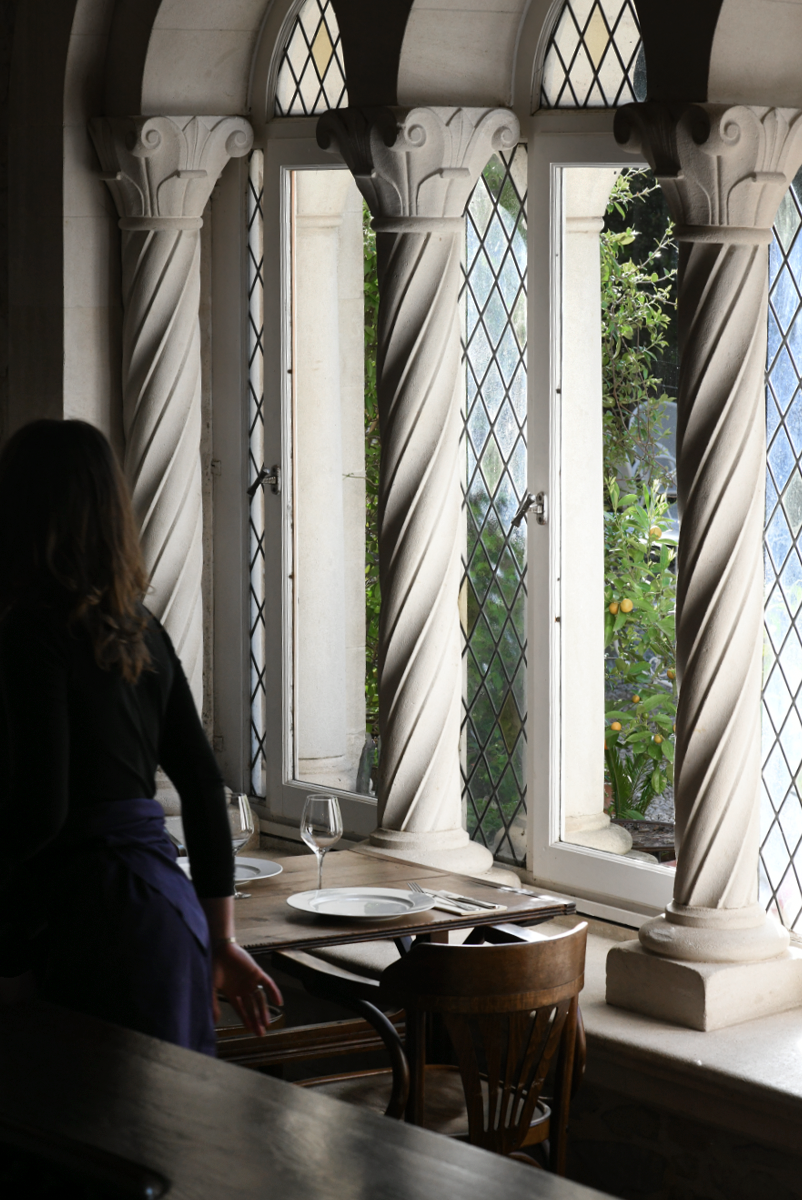 Un serveur se tenant près d'une table dans une salle avec de grandes fenêtres ornées de colonnes torsadées et de fenêtres en treillis. C'est l'intérieur du restaurant La Courtille