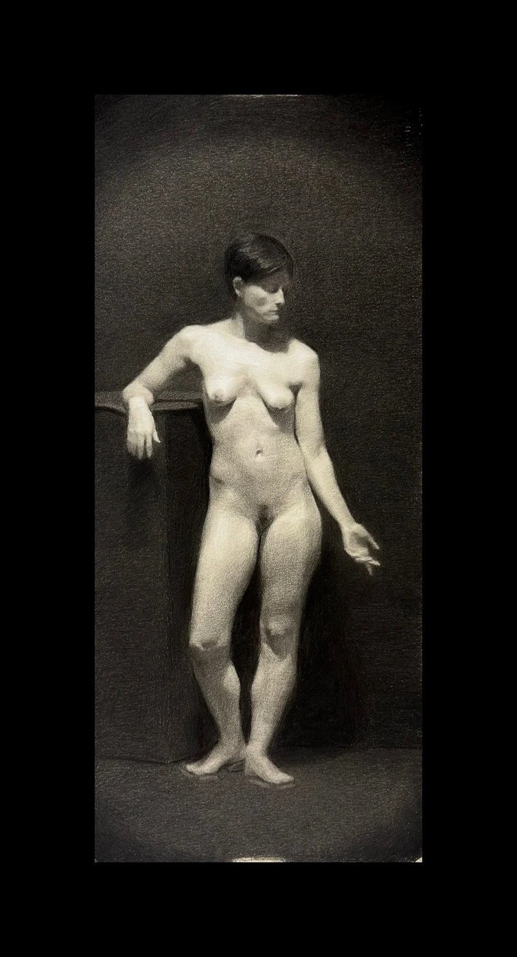 A black and white photo of a nude woman with short dark hair, standing against a dark background, leaning on a surface with her left arm, looking down.
