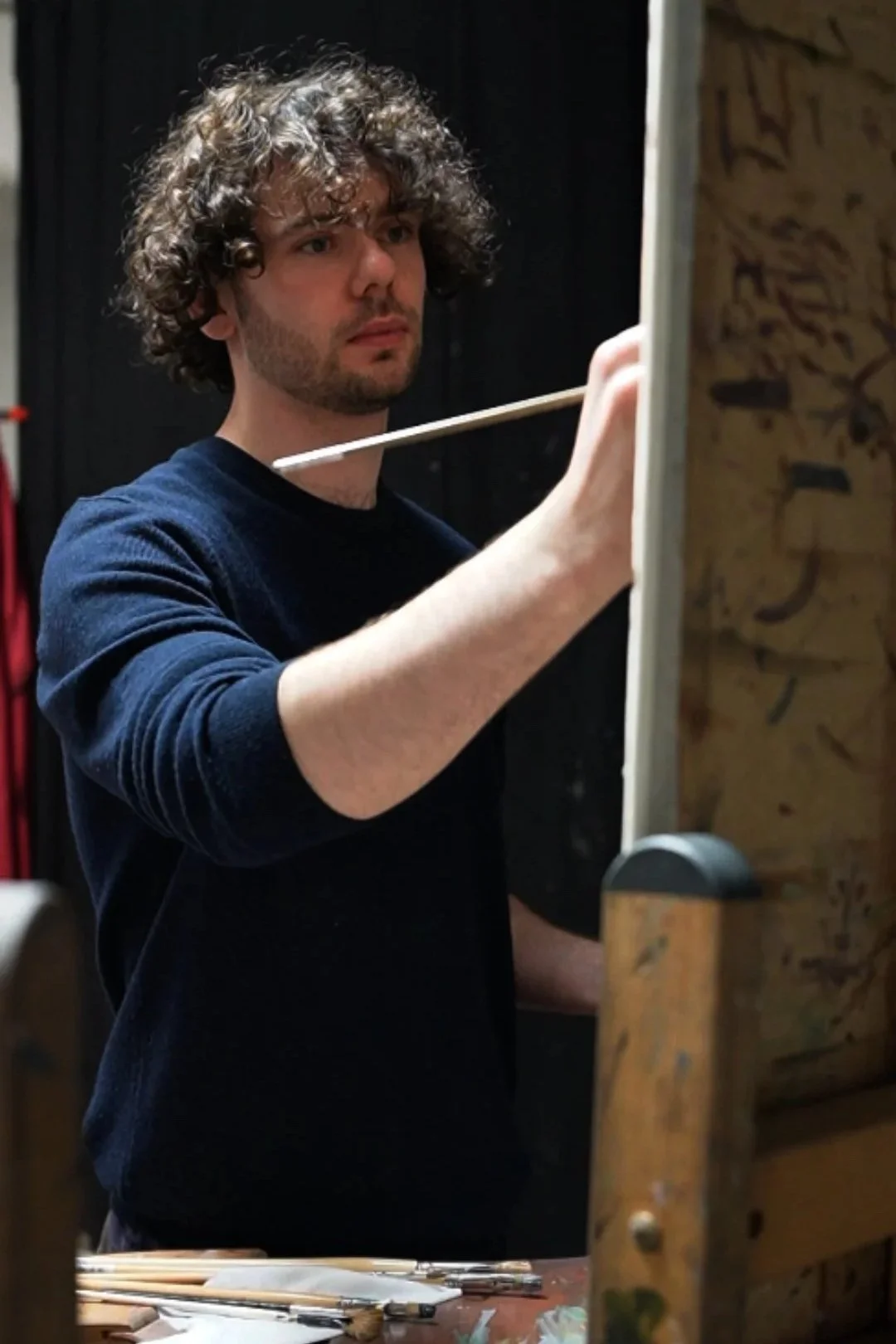 A man with curly hair, wearing a navy blue sweater, is painting on a canvas in a studio.