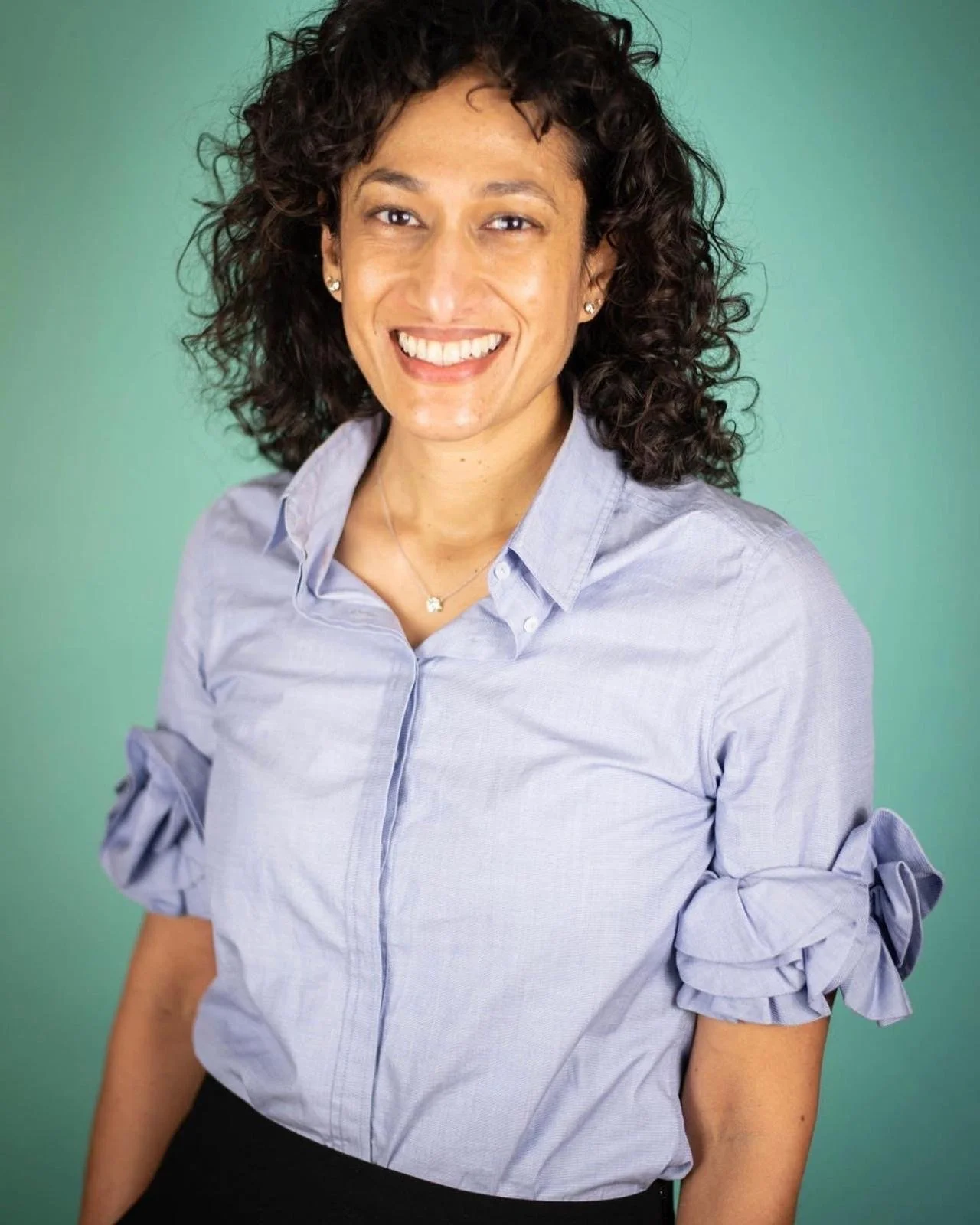 A woman with curly dark hair smiling, wearing a light blue blouse with rolled-up sleeves and a necklace, standing against a green background.