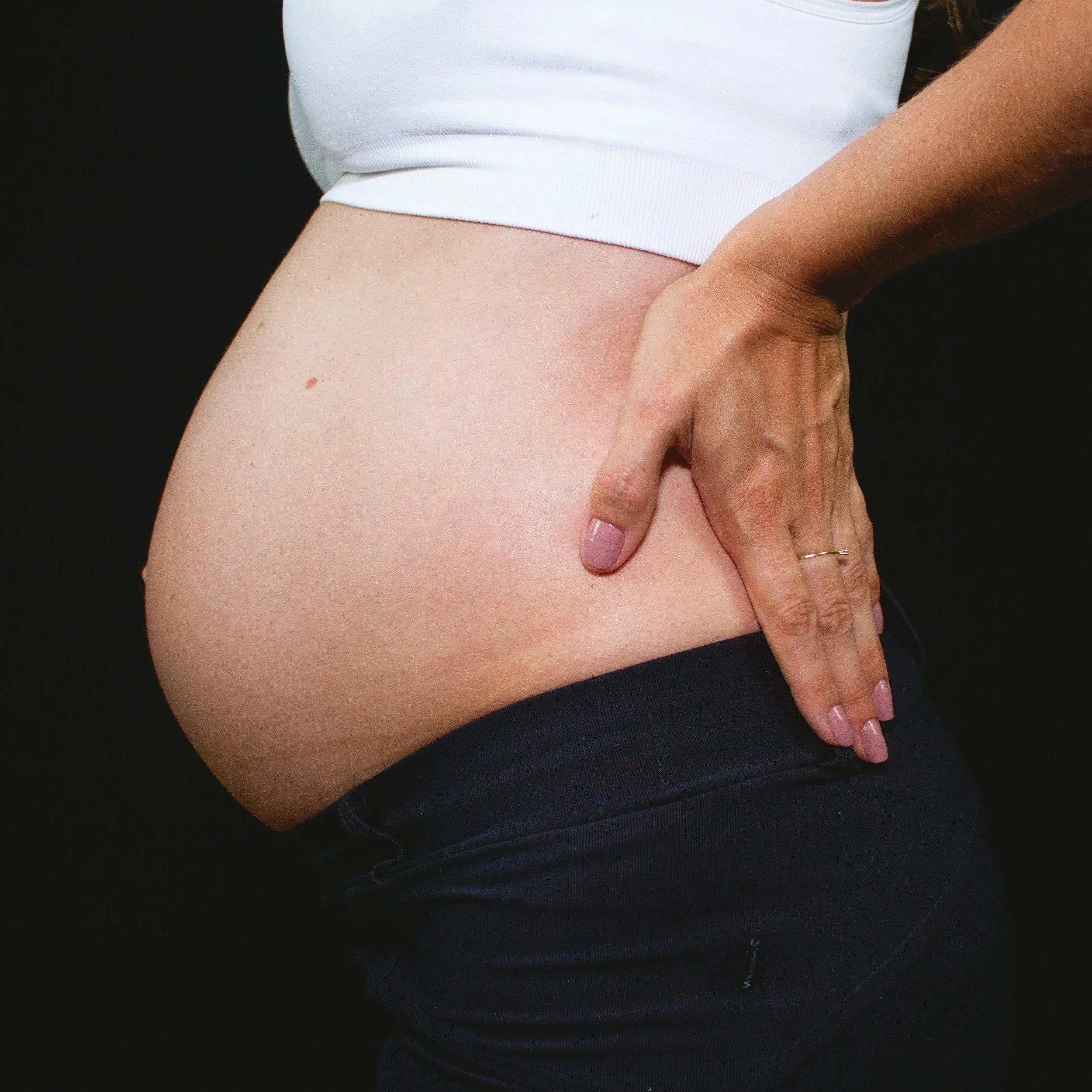 Close-up of a woman's waist with a hand resting on her hip, wearing a white top and dark jeans against a black background.
