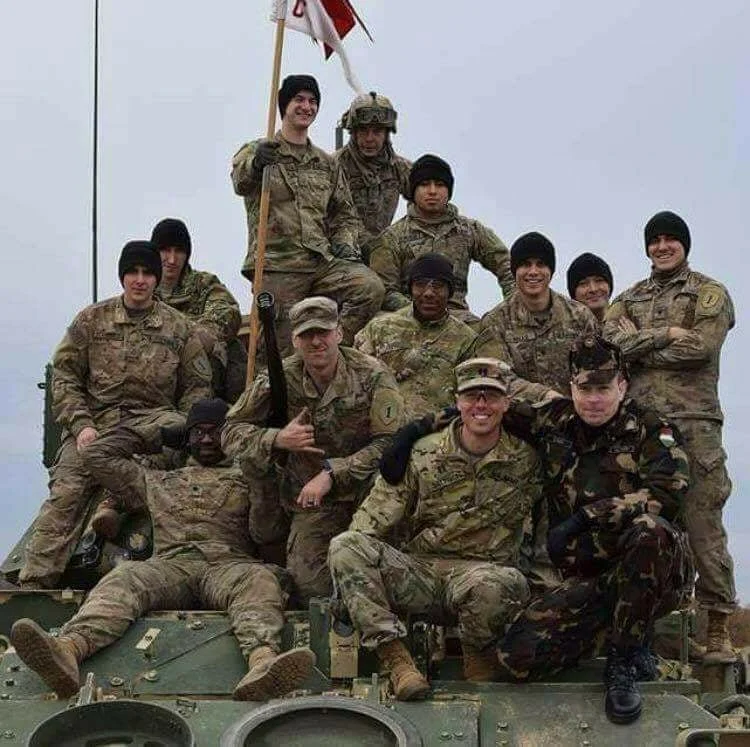 Group of soldiers in uniform posing on top of a military vehicle, with some standing, sitting, and lying down, and one holding a flag.