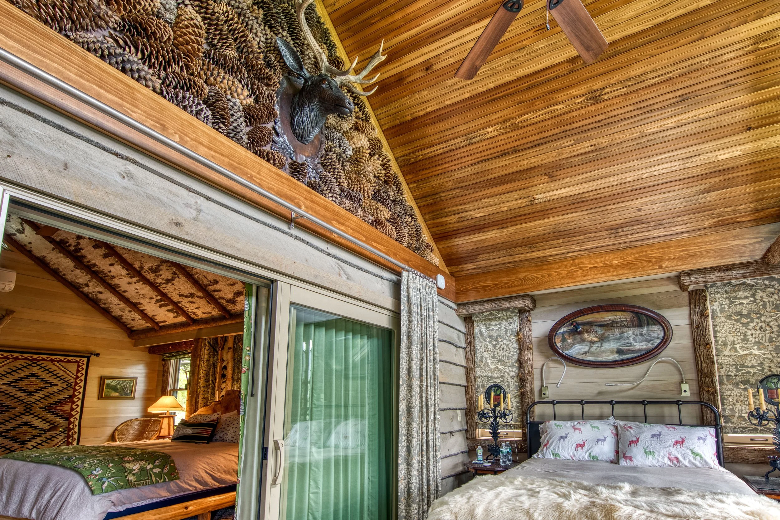 A rustic bedroom with wooden walls and ceiling, a mounted deer head with antlers above pinecone decorations, a metal bed with deer-patterned pillows, and wall-mounted lamps.