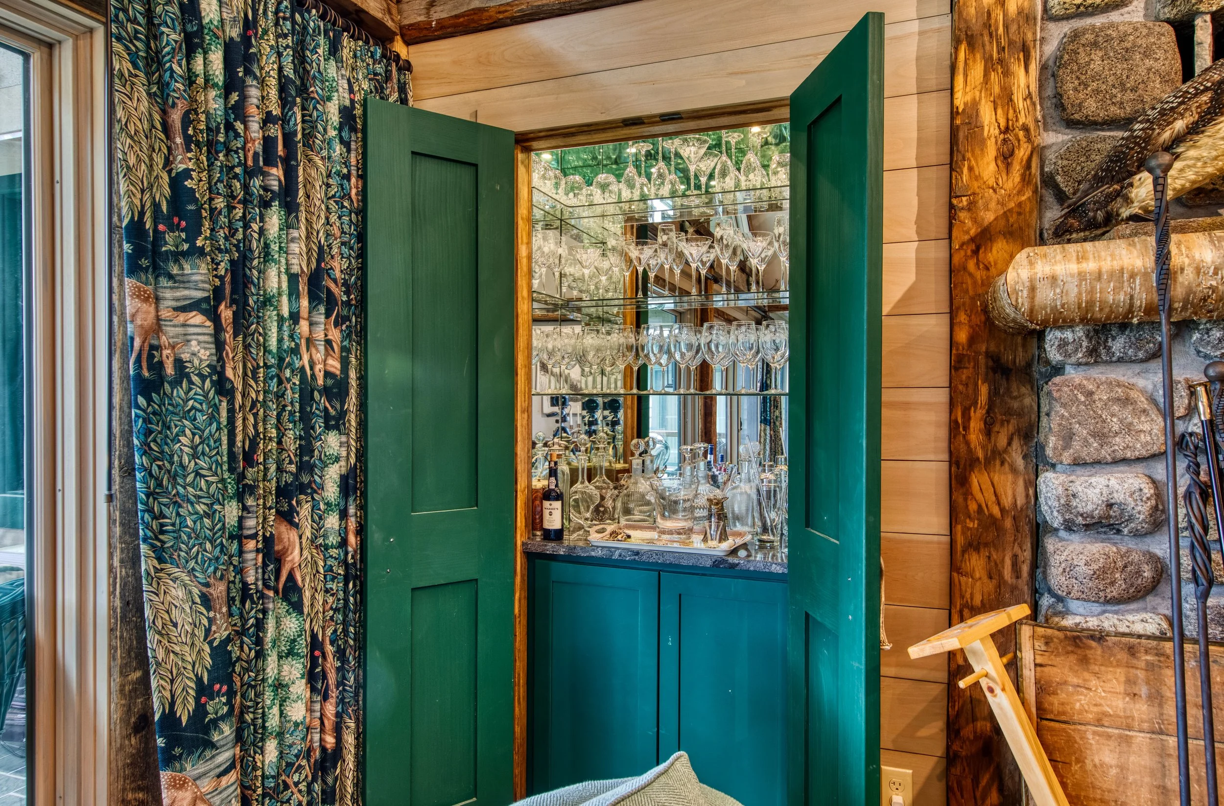 A cozy room with a display of glassware inside a green cabinet with open doors, next to a wall with patterned curtains and a stone and wood wall on the other side.
