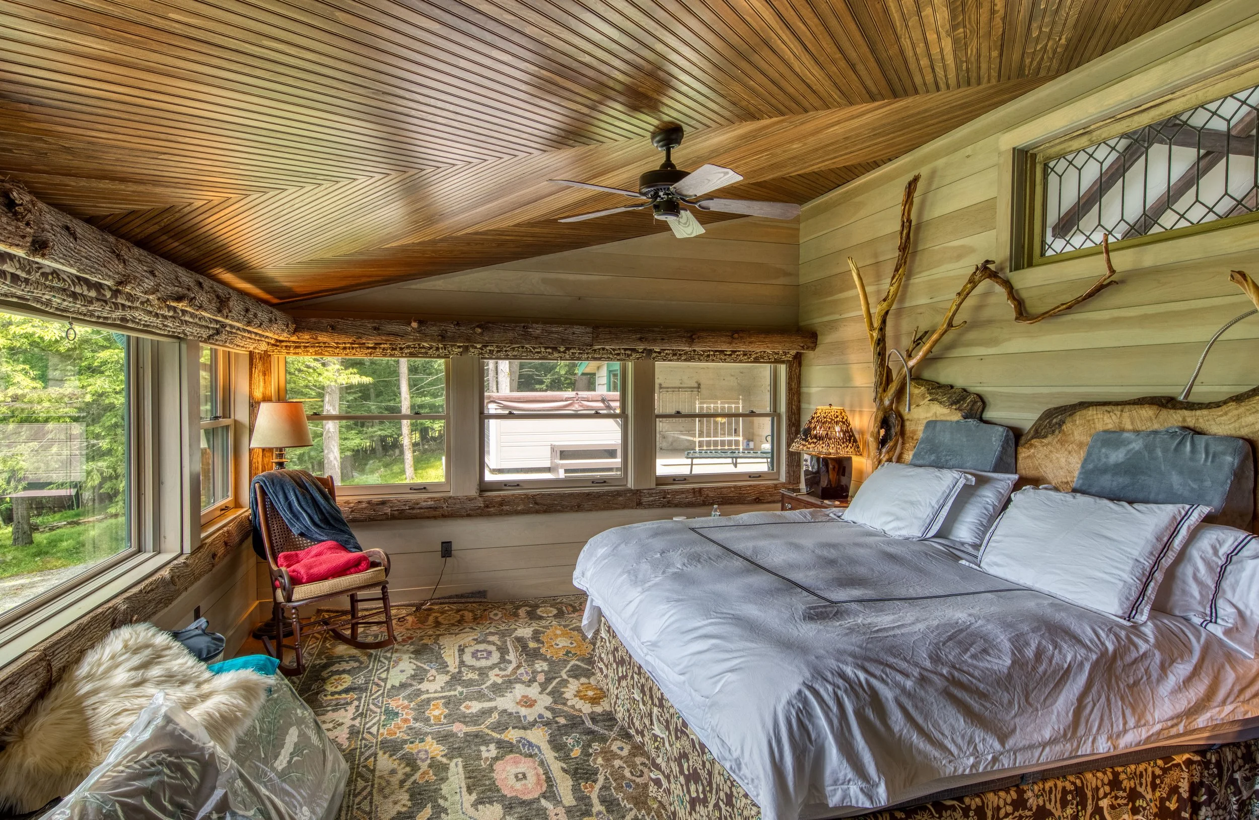 Cozy bedroom with wood-paneled ceiling and walls, large windows overlooking greenery, bed with a rustic wooden headboard and white bedding, a rocking chair with blanket, a lamp, and a ceiling fan.