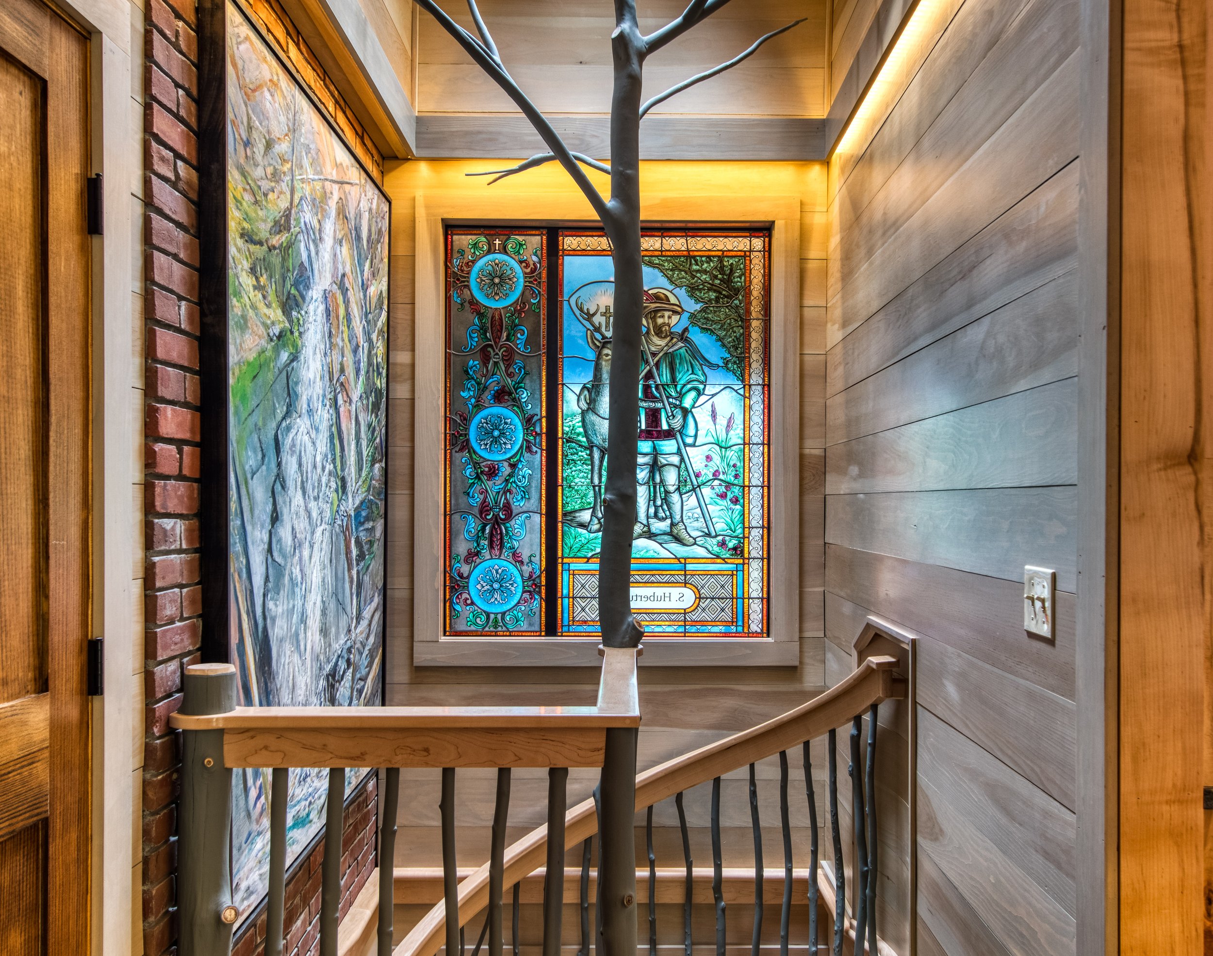 A staircase with a wooden handrail and black iron spindles, a stained glass window depicting a shepherd with a staff and a sheep, a large painting on the left wall, and wooden walls.