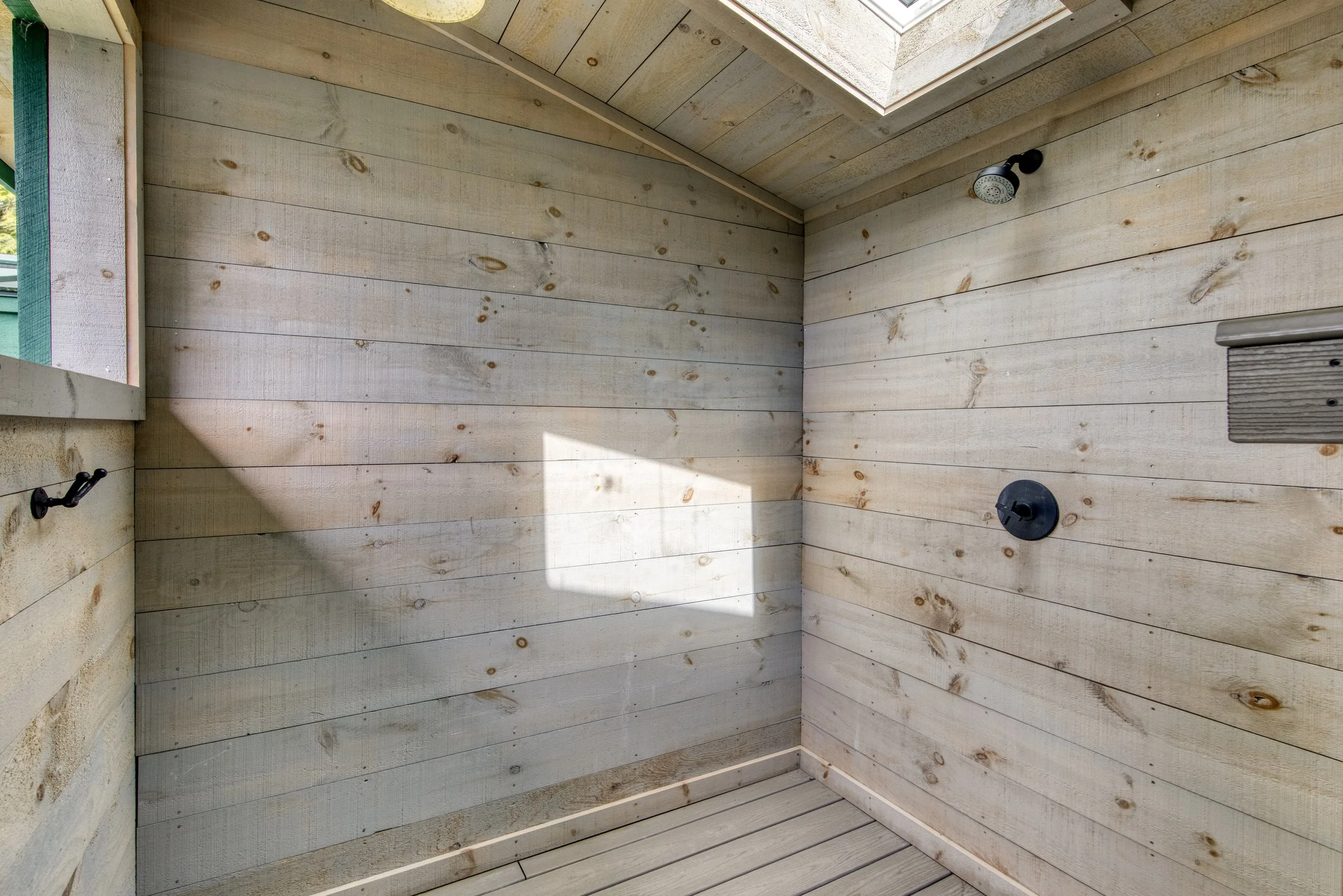 Outdoor shower with wooden walls and floor, black showerhead, and control knob, with a small window and skylight above.