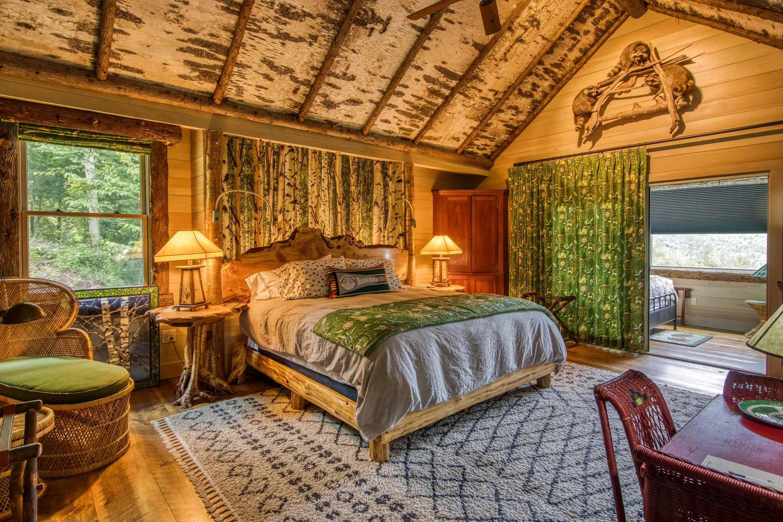 A rustic cabin bedroom with a wooden bed, two lamps, a wardrobe, green patterned curtains, a window showing a green outdoor view, and decorative raccoon and tree branch artwork on the wall.