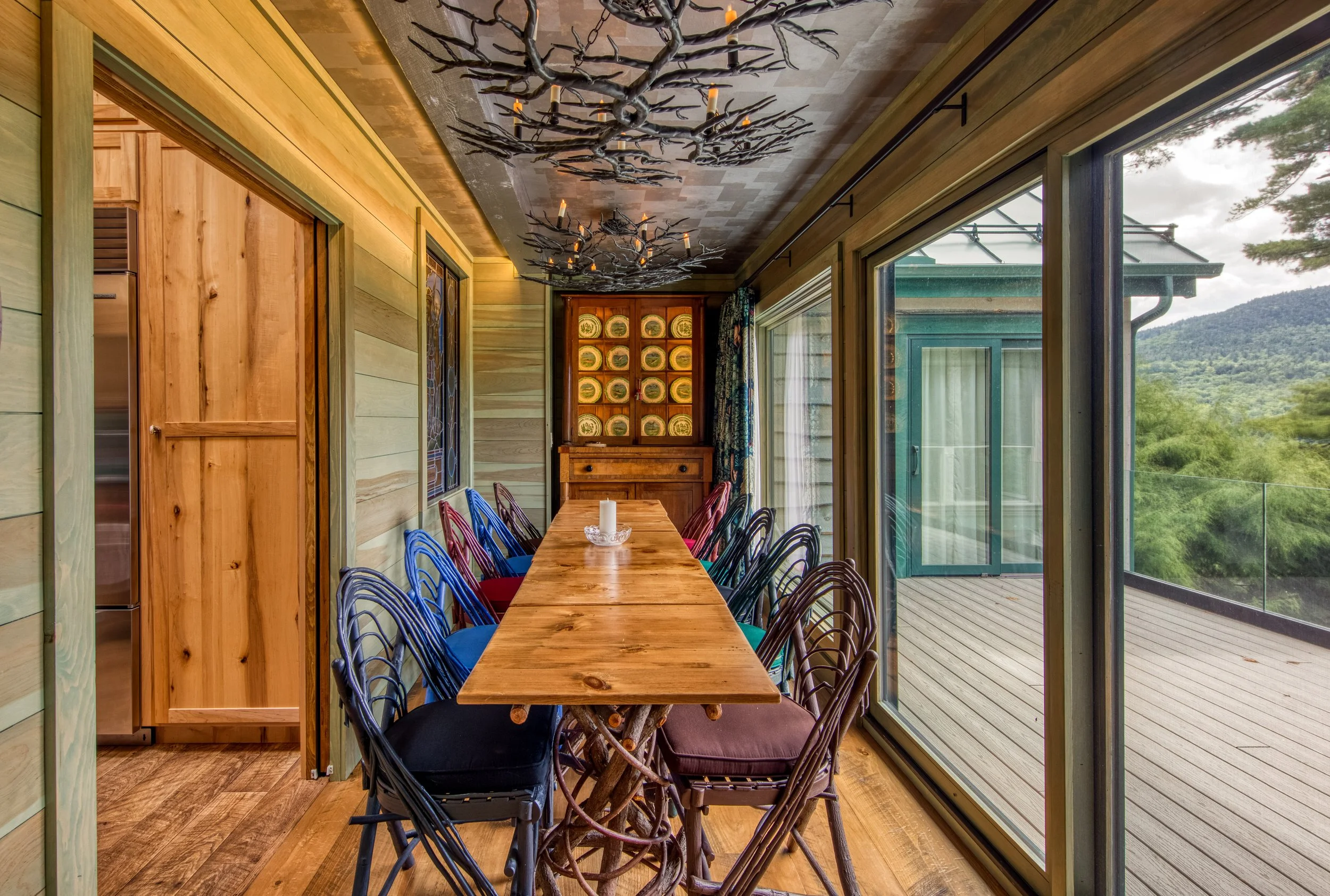 Dining room with a long wooden table surrounded by multicolored chairs, large glass sliding doors leading to a balcony with scenic mountain views, wooden wall paneling, and decorative black chandeliers resembling tree branches with candles.