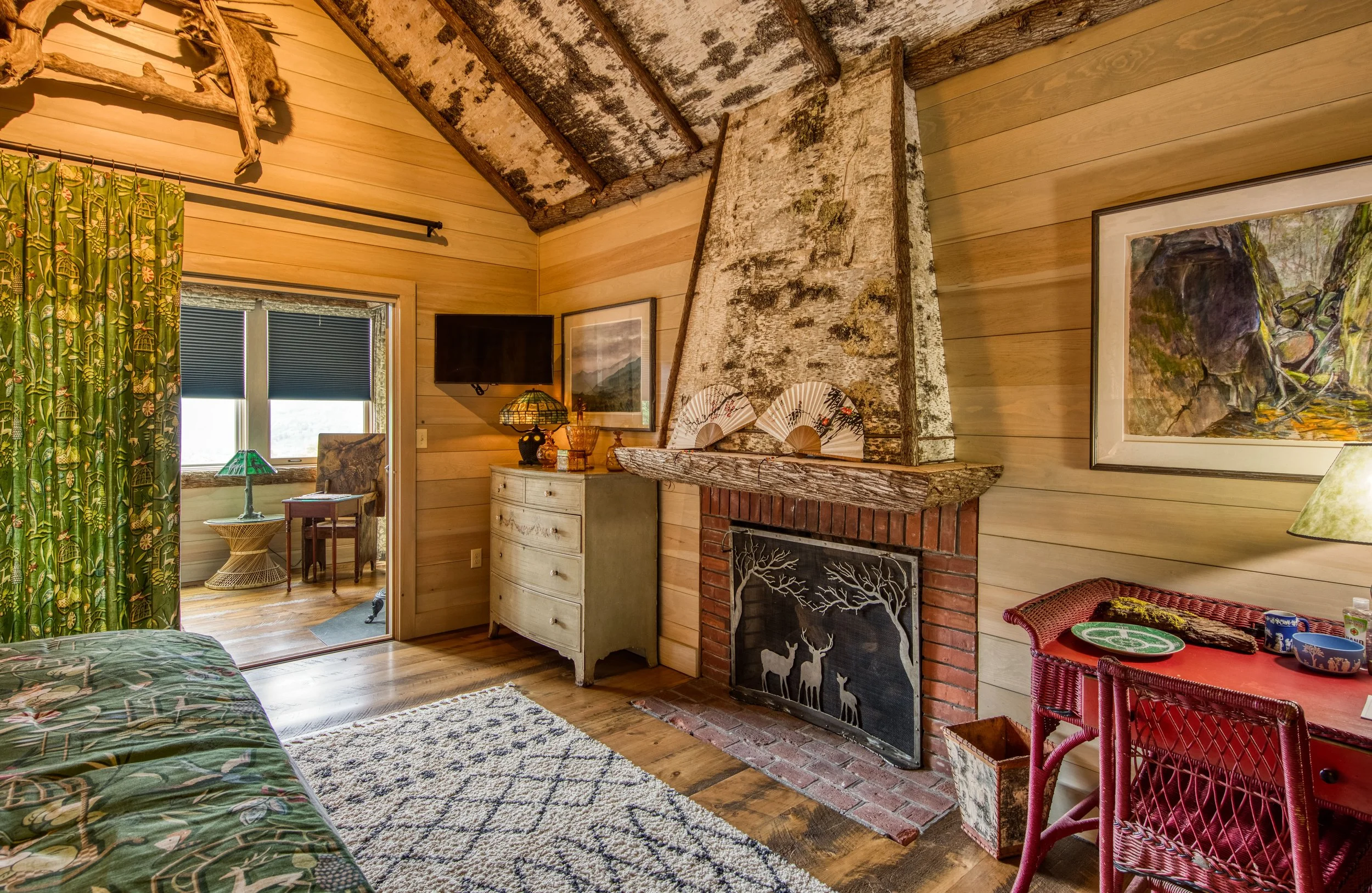Cozy cabin living room with wood-paneled walls, a rustic stone and brick fireplace with deer silhouettes, a window with green patterned curtains, an area rug, a red wicker table with dishes, and a small sitting area visible through a doorway.