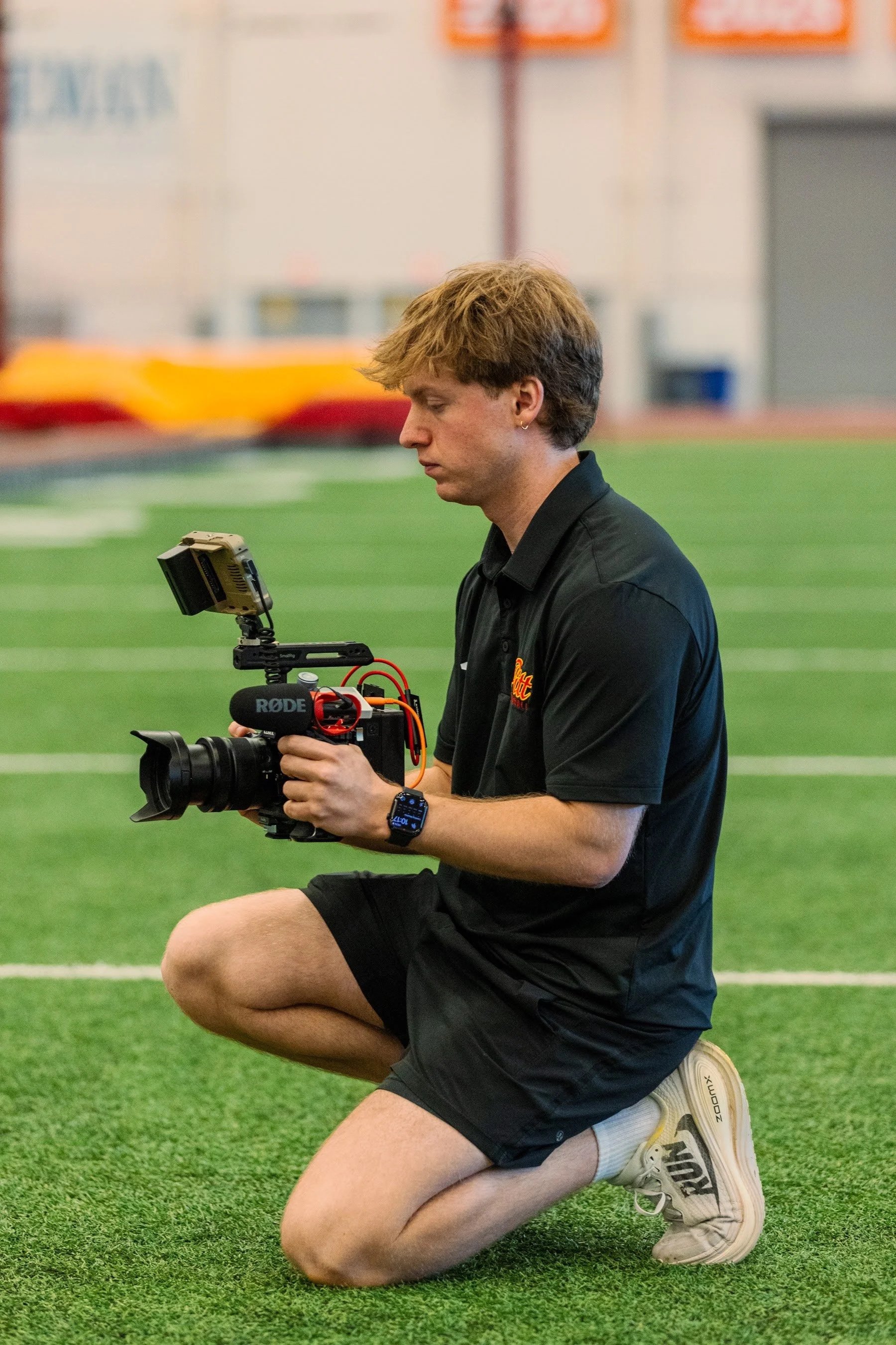 A man kneeling on a sports field holding a professional video camera, wearing a black shirt, black shorts, and white sneakers.