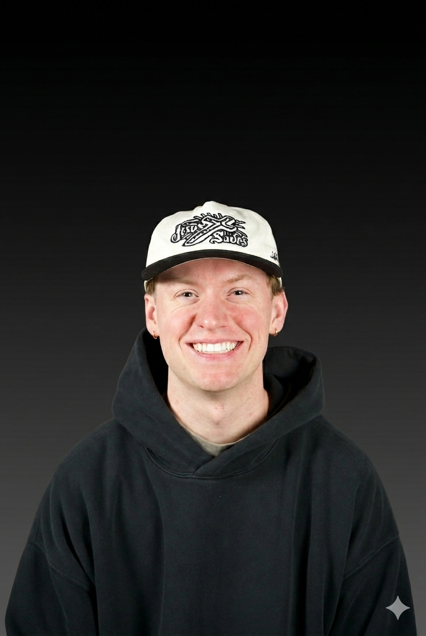 Young man smiling, wearing a black hoodie and a white baseball cap with a black brim and black embroidery, against a black background.