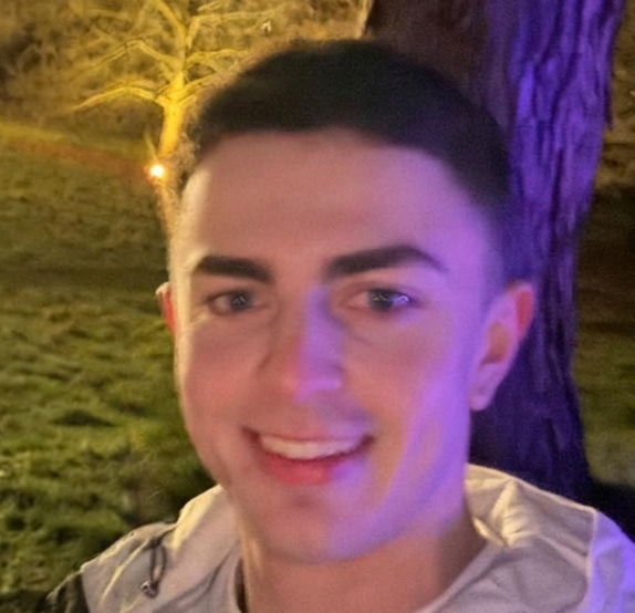 A young man smiling outdoors at night, standing near a tree, with purple and yellow lighting in the background.