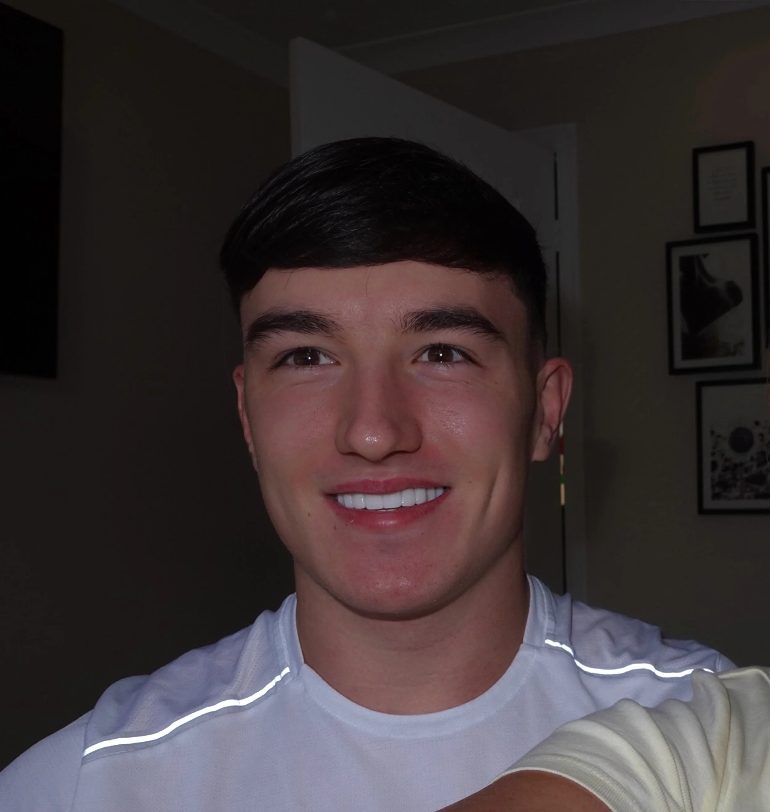 Close-up of a young man's face with dark hair, wearing a white shirt, smiling, in a room with framed pictures on the wall.