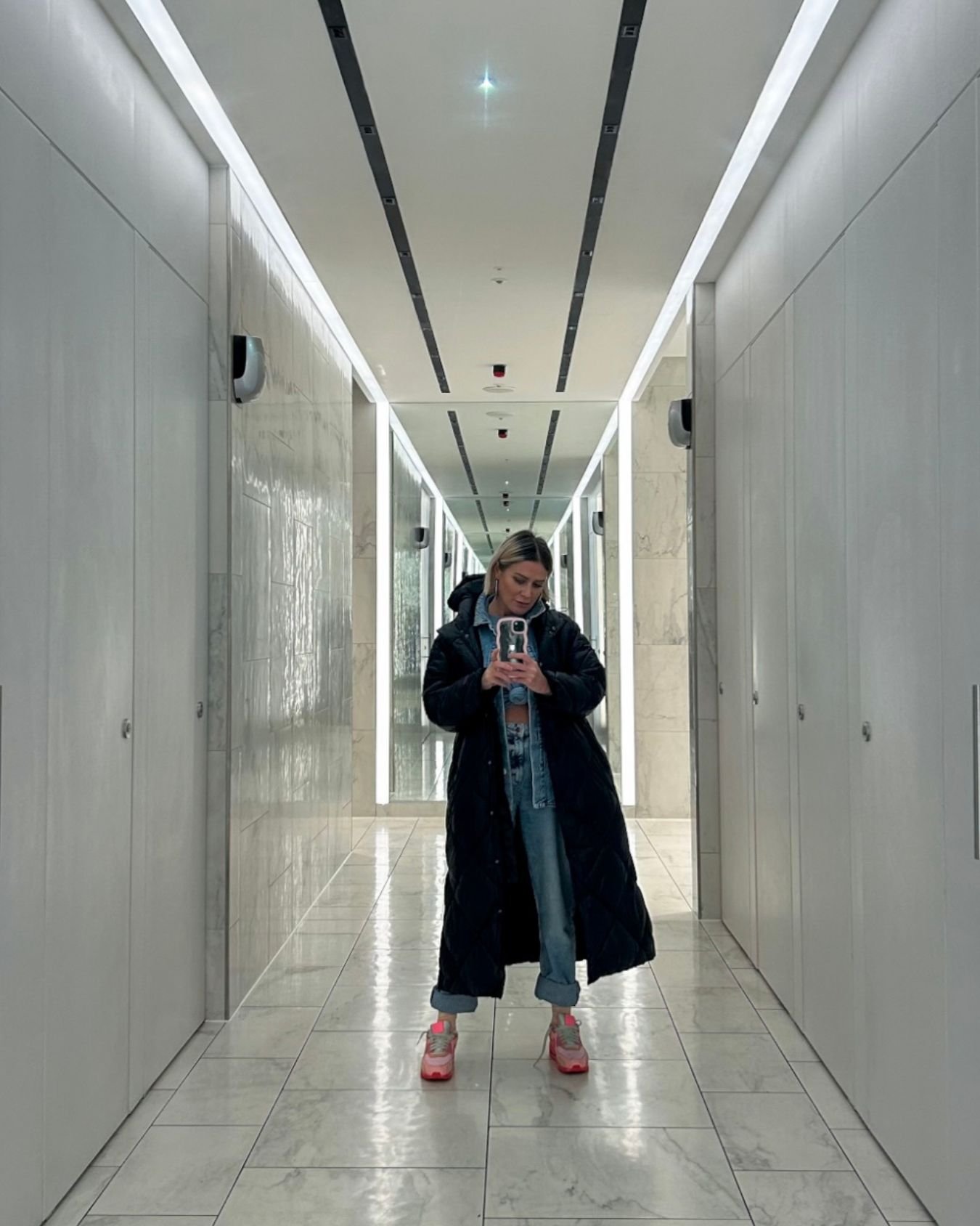 A woman in a long black coat is taking a selfie on the mirror.