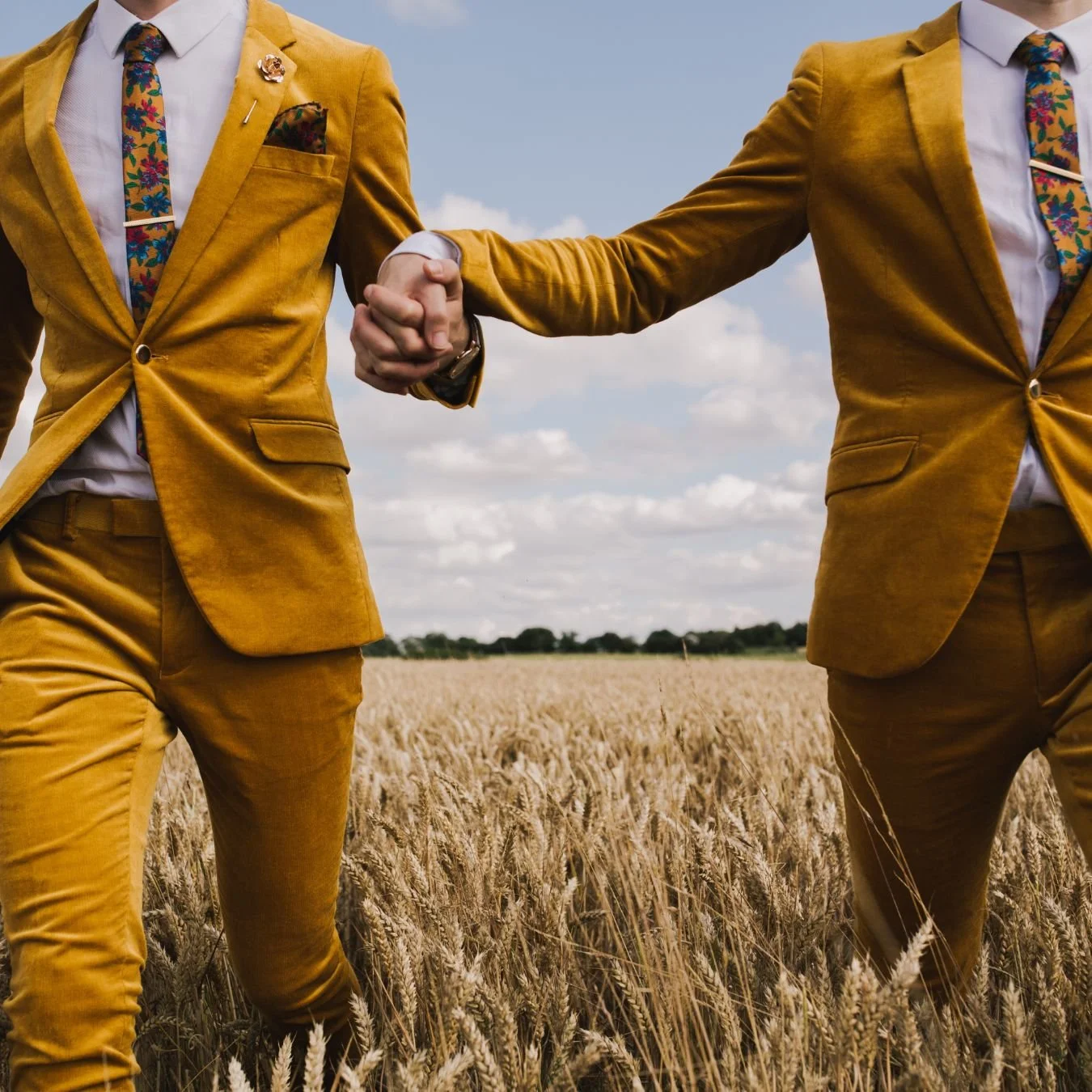 2 grooms wearing mustard suits are holding hands in a cornfield.