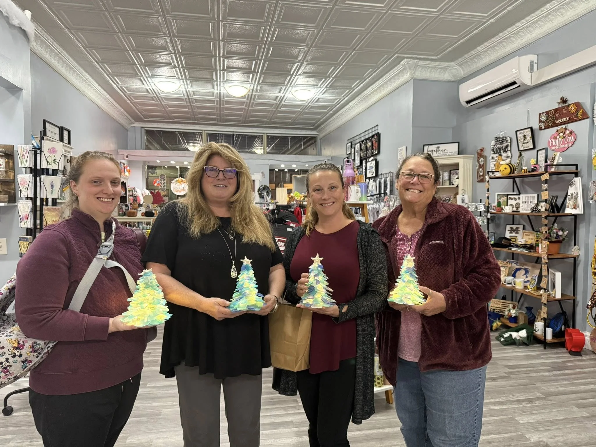Four women standing inside a gift shop, each holding a colorful ceramic Christmas tree. They are smiling and posing for the photo. The shop has Christmas decor and items on shelves in the background.