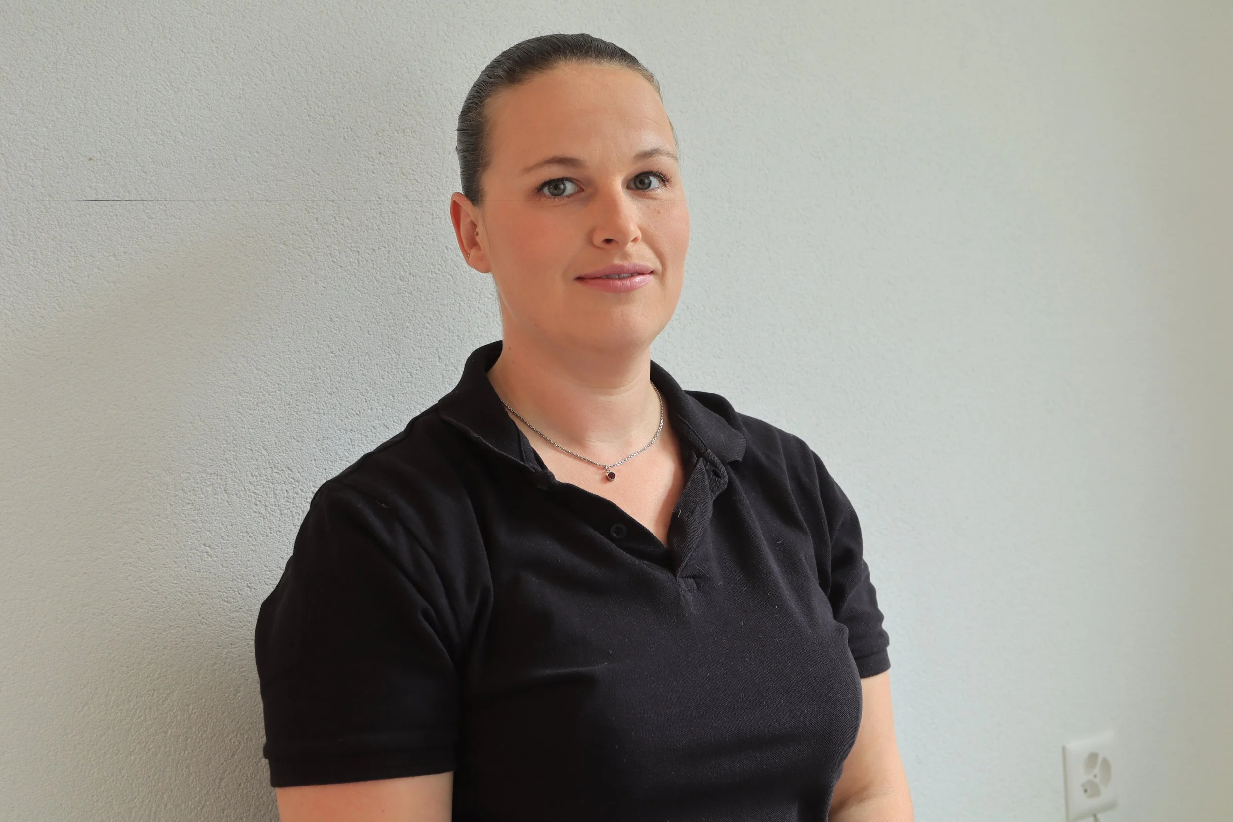 Eine Frau mit braunen Haaren, die zurückgebunden sind, trägt ein schwarzes Polo-Shirt und eine Halskette, steht vor einer weißen Wand.
