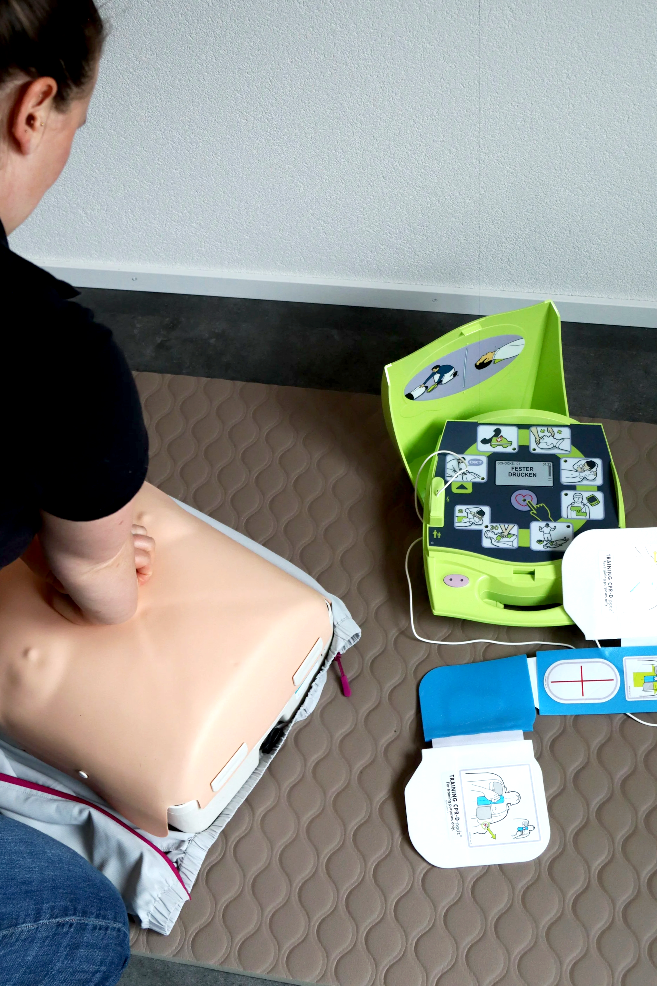 Lebensrettungsübung mit einem Dummy und einem Automatisierten Externen Defibrillator auf einem Boden. Die Übung findet wahrscheinlich in einer Schulungsumgebung statt.