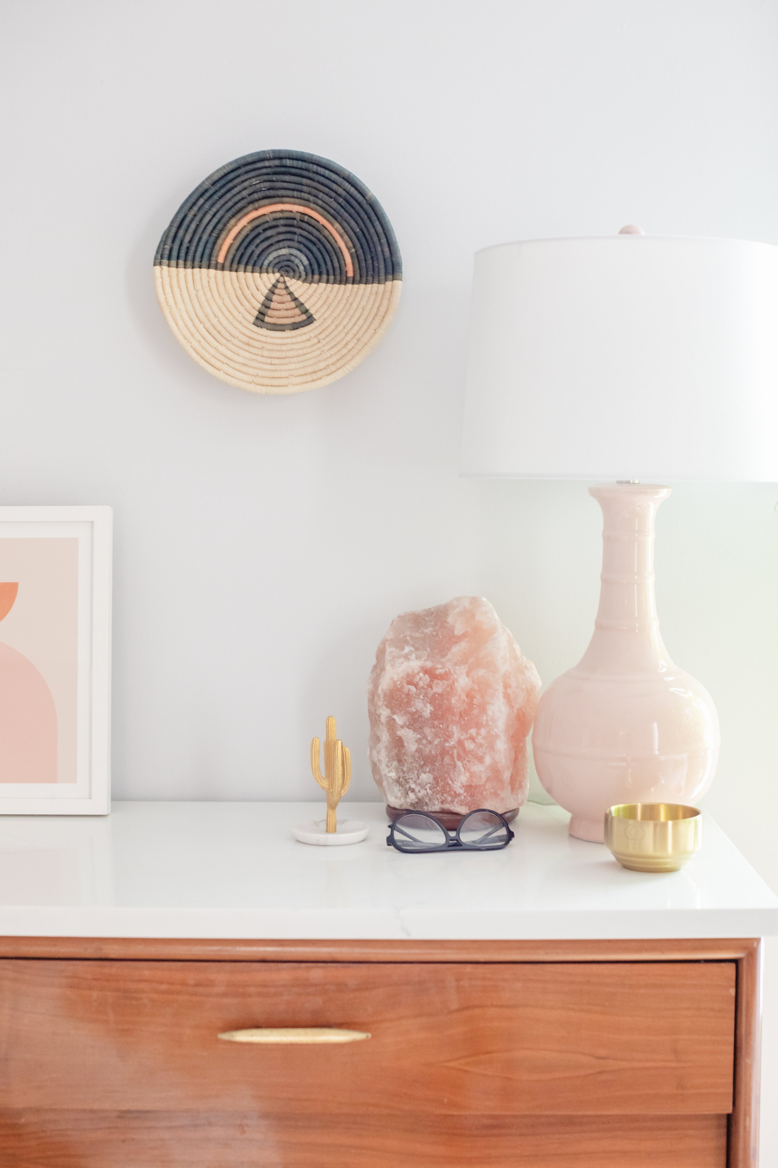 A white dresser with wooden drawers, topped with decorative objects including a pink salt lamp, a gold bowl, a small cactus figurine, a pair of glasses, and a pink vase. There is a framed abstract artwork on the left, and a round woven wall hanging with a geometric pattern and rainbow colors on the wall above.