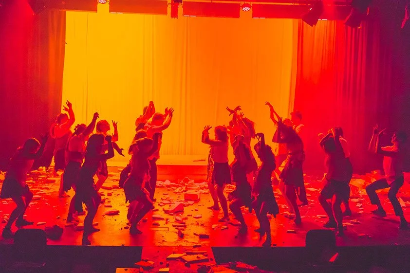 Photo from the production 'The Howl'; a group of performers dances energetically on a dimly lit stage with a vibrant orange-yellow backdrop, surrounded by scattered papers. Their dramatic poses create lively silhouettes.