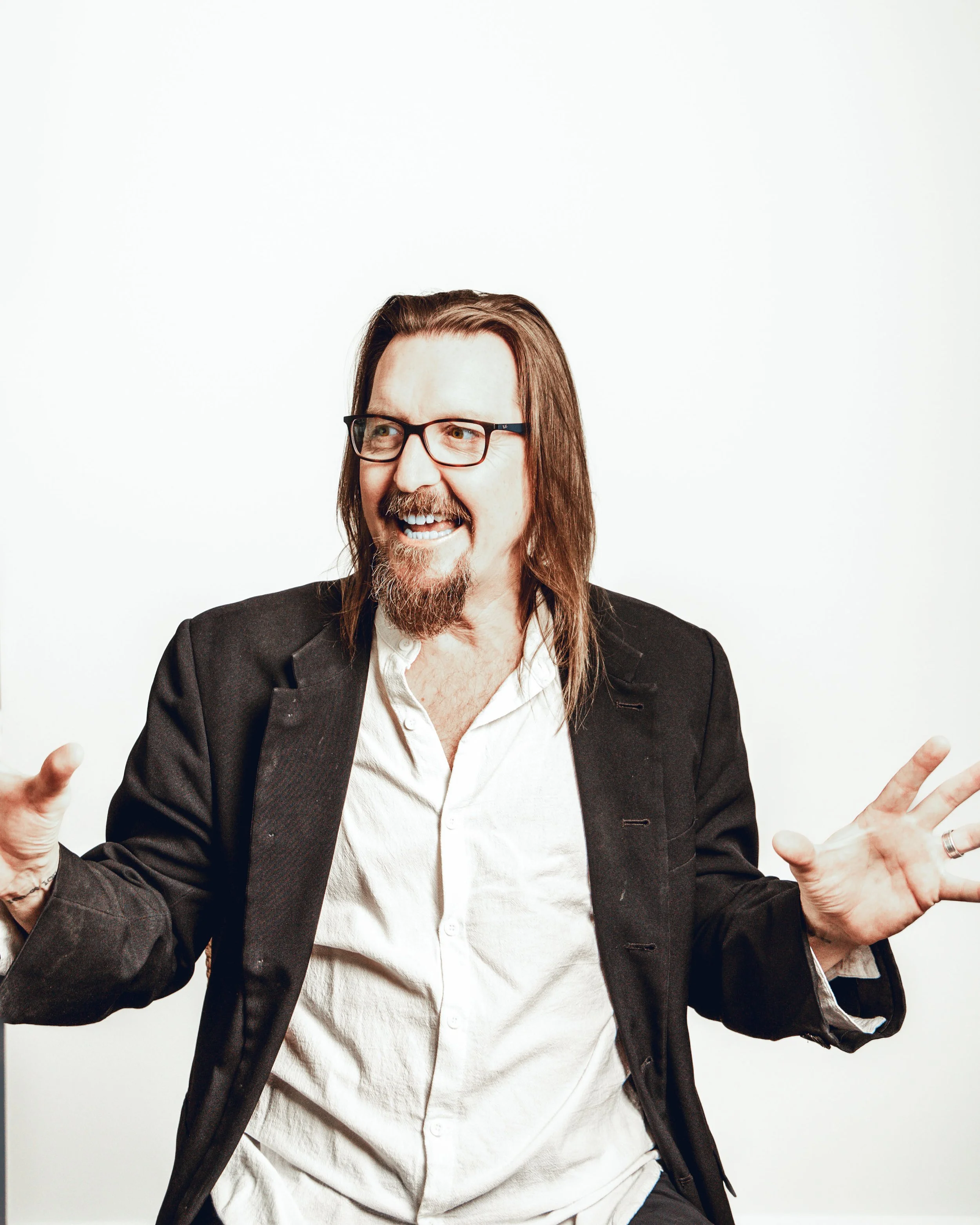 Simon Tate with long hair, glasses, and a beard smiles and gestures with his hands. He is wearing a white shirt and a black blazer, standing against a plain white background.