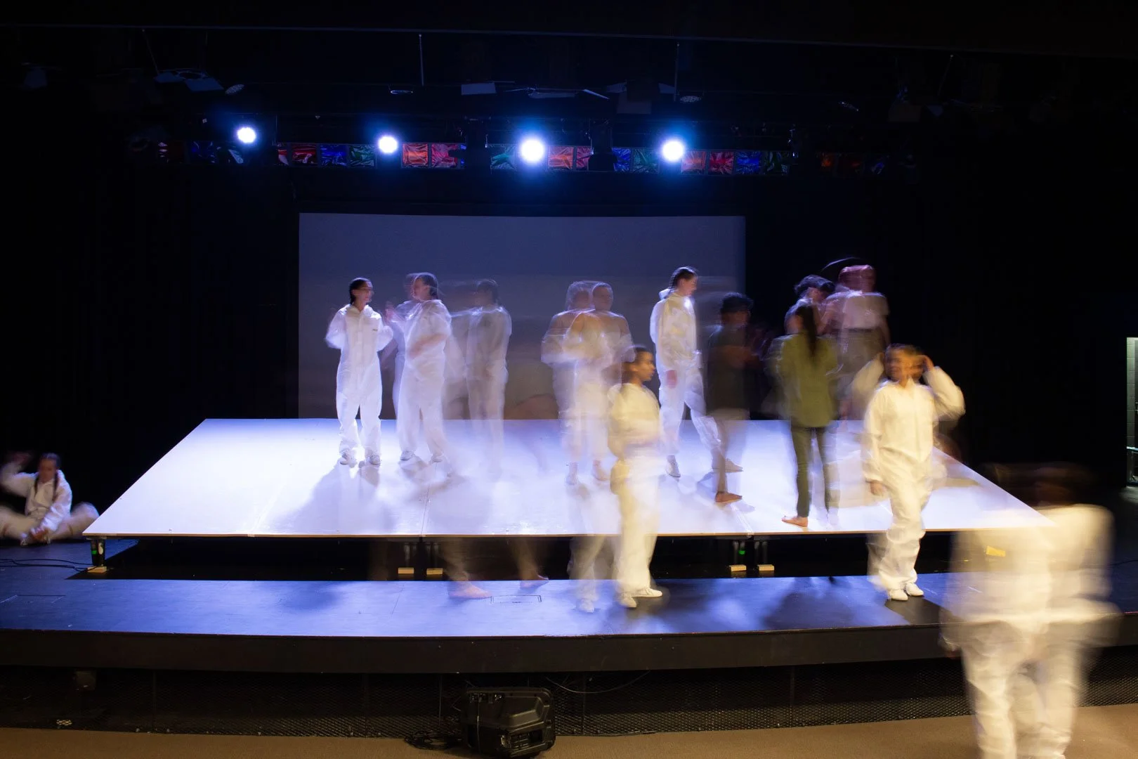 Photo from the production 'The Ark'; people on stage performing with some wearing white costumes, in a dark theater with stage lights overhead.
