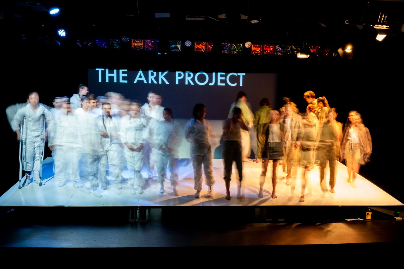 Photo from the production 'The Ark'; a stage with performers in motion, some appearing blurred, under a sign that says "The Ark Project."
