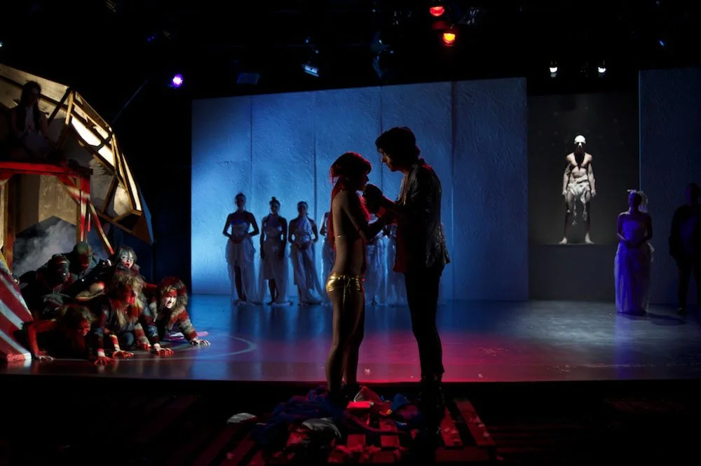 Theater scene with people in costumes, including a woman receiving something from a man, while others dressed as animals are on the stage and in the background.