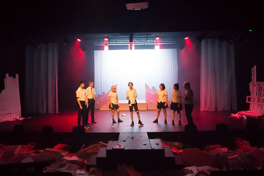 Seven people in school uniforms stand on a dimly lit stage with a red and purple glow from the production 'The Howl'..