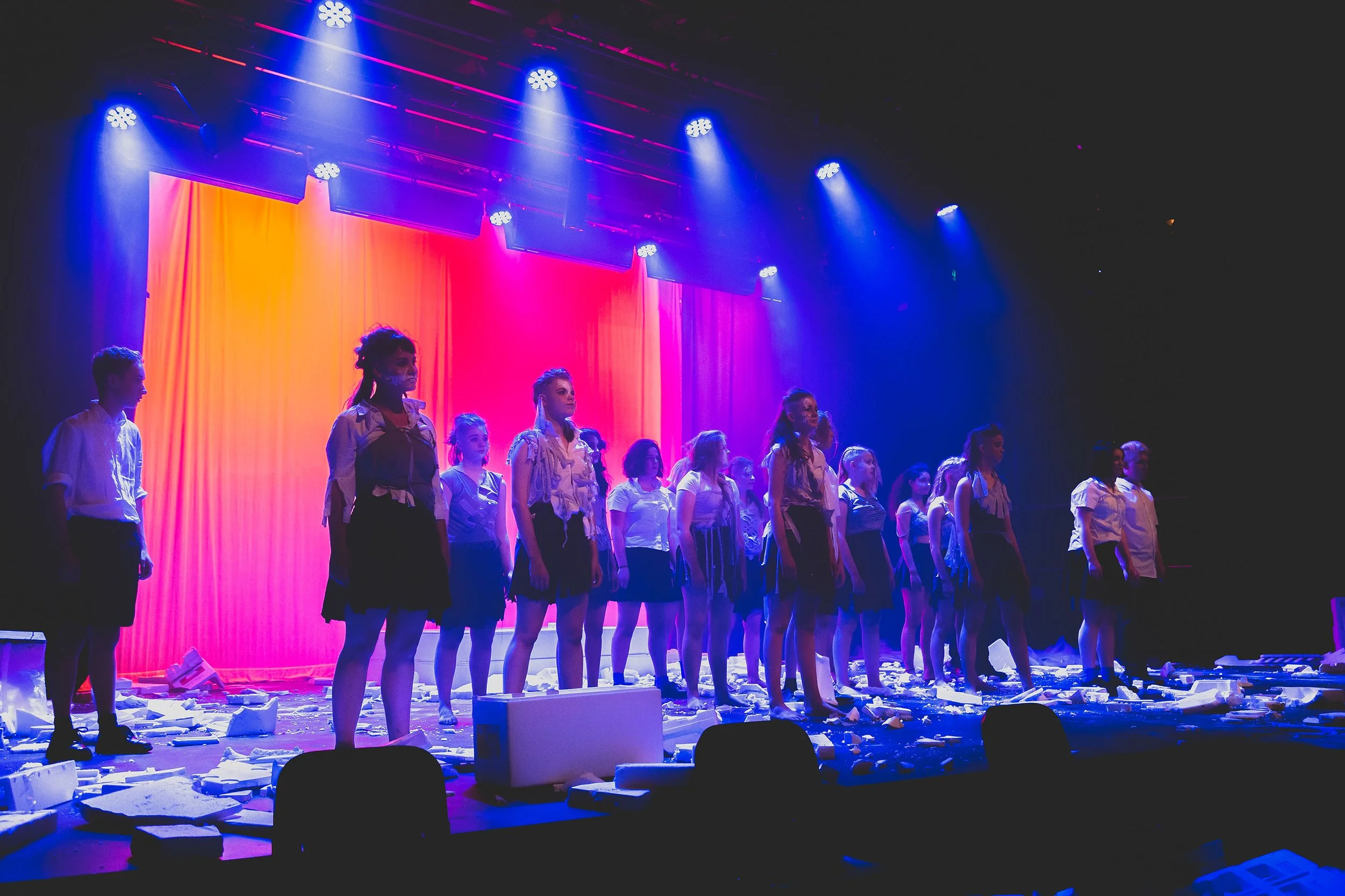 Photo from the production 'The Howl'; a group of young performers in school uniforms stand in a line on a stage, lit by blue and pink lights, with scattered papers and debris on the floor creating a dramatic scene.
