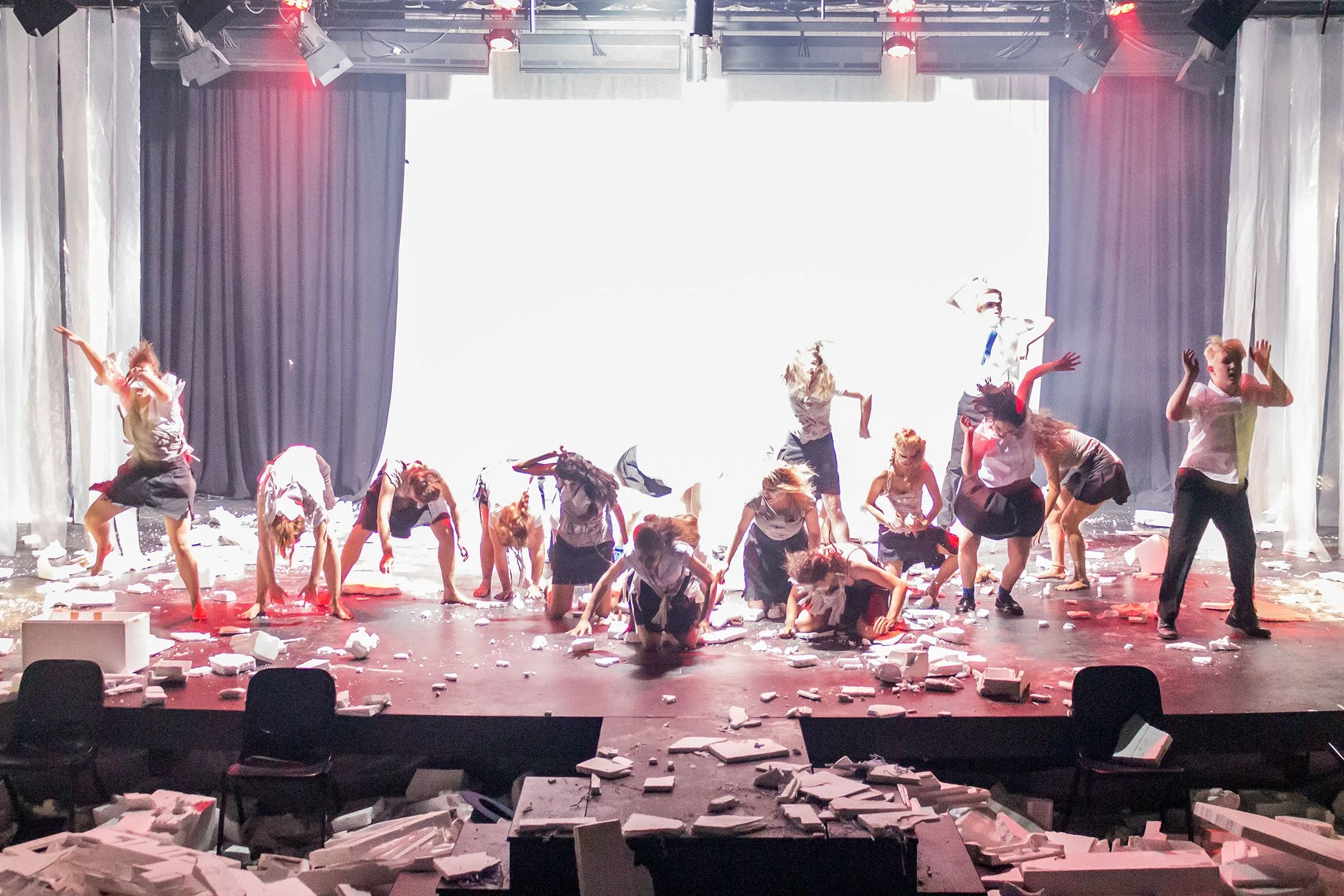 Photo from the production 'The Howl'; a group of performers on a stage strike dynamic poses amid scattered debris and dramatic lighting, with some kneeling or crouching and others standing.