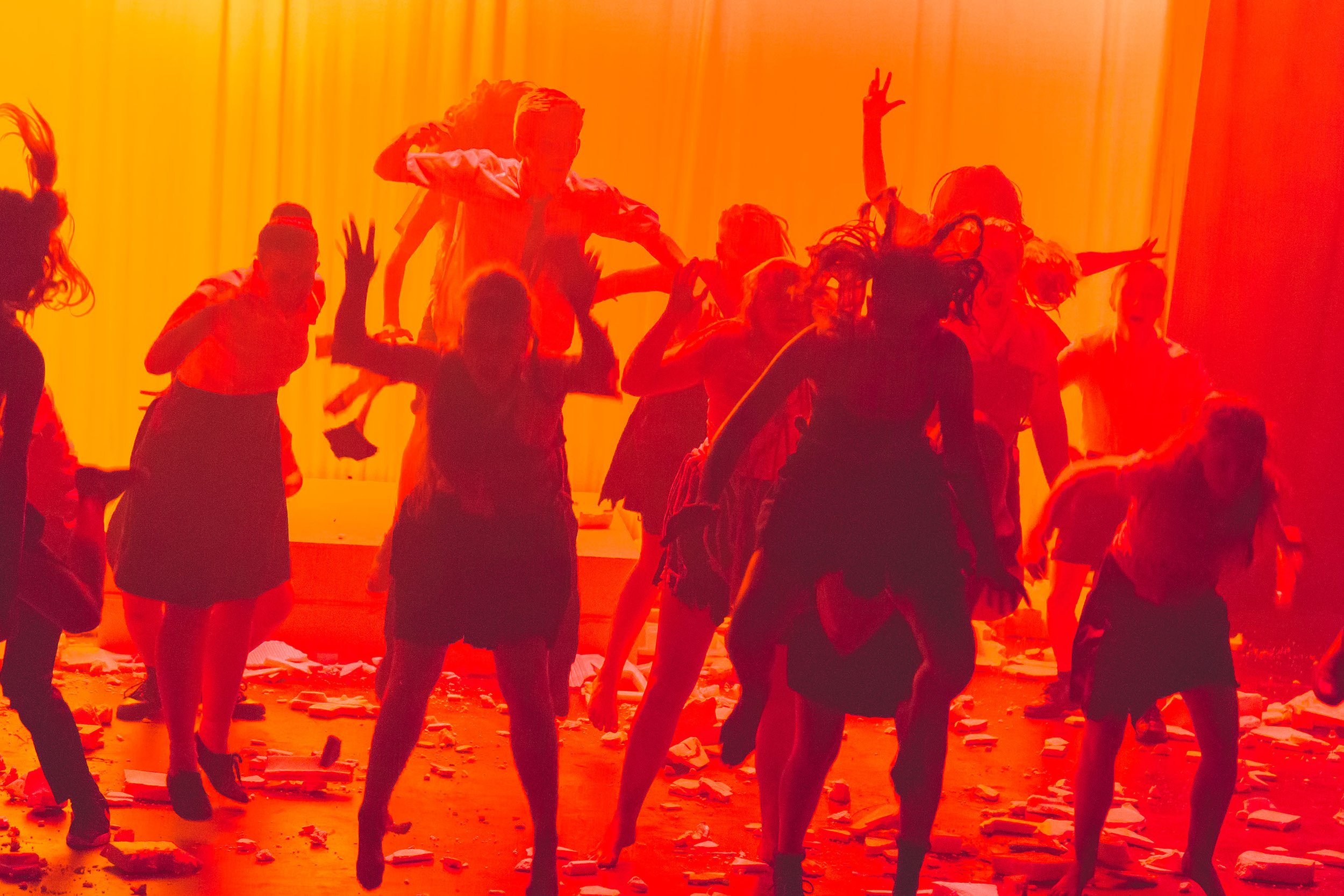 Photo from the production 'The Howl'; a group of people performing in a room illuminated with warm orange light. The scene includes scattered debris on the floor and an energetic atmosphere.
