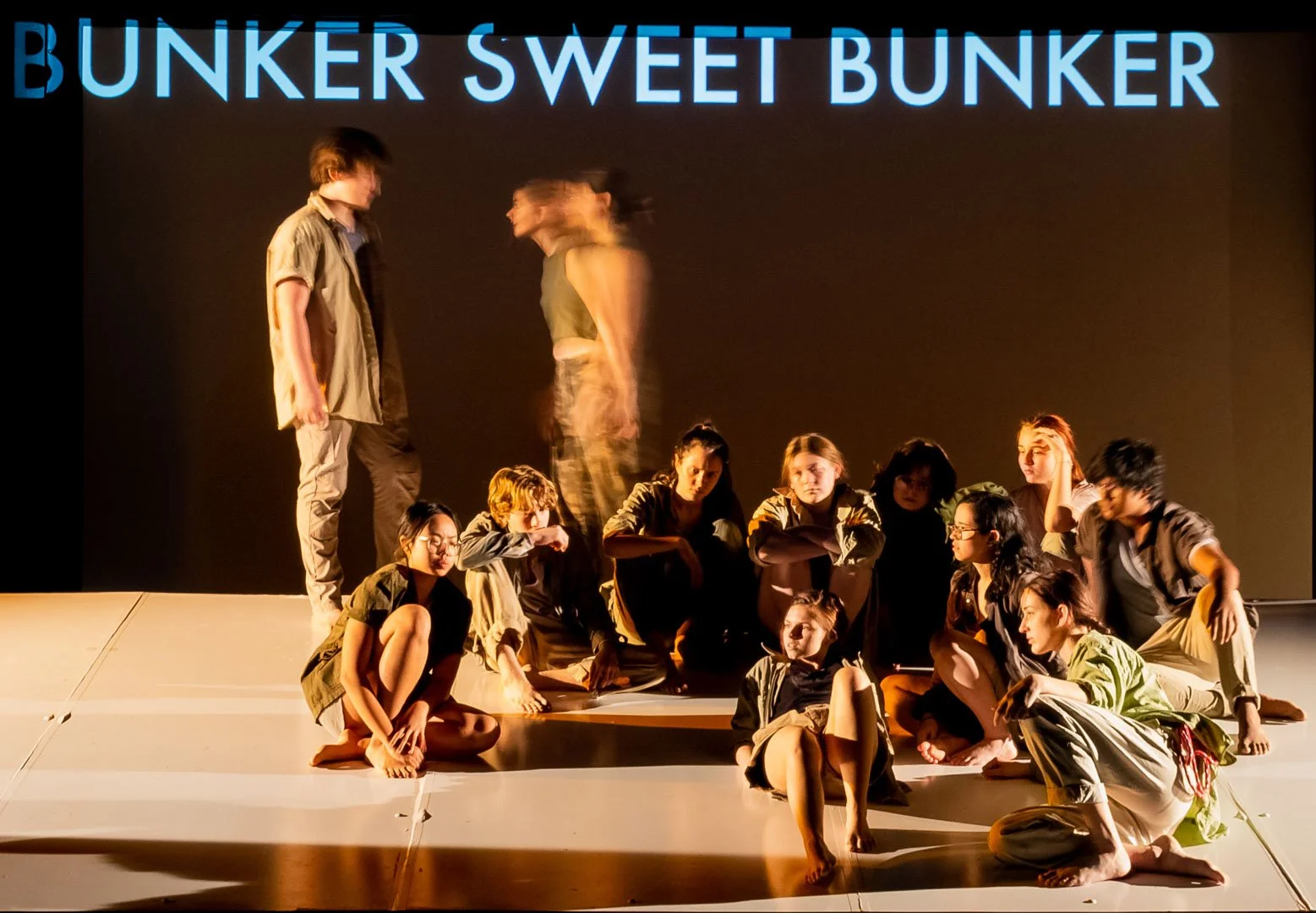 Photo from the production 'The Ark'; a group of young people on stage performing, with two standing figures at the front and others seated or crouched behind them.