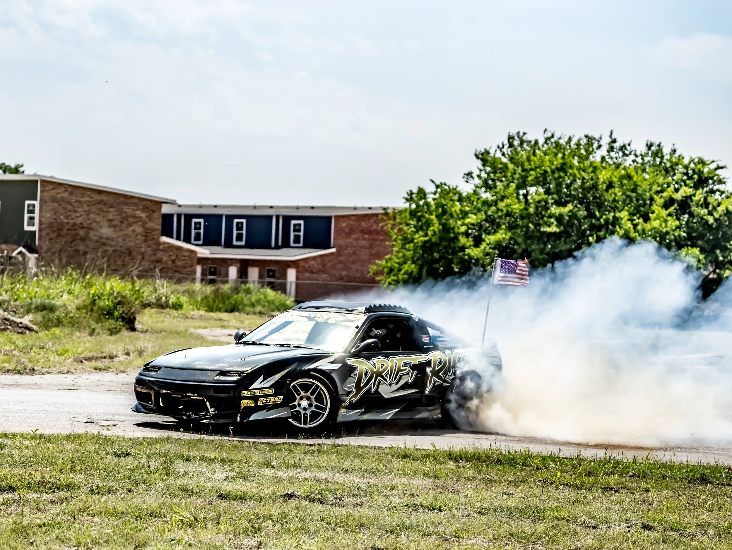 Black sports car performing a burnout with smoke, tinted windows, and racing decals, including the word 'Drifter', on a street with a house, trees, and a partially visible American flag in the background.