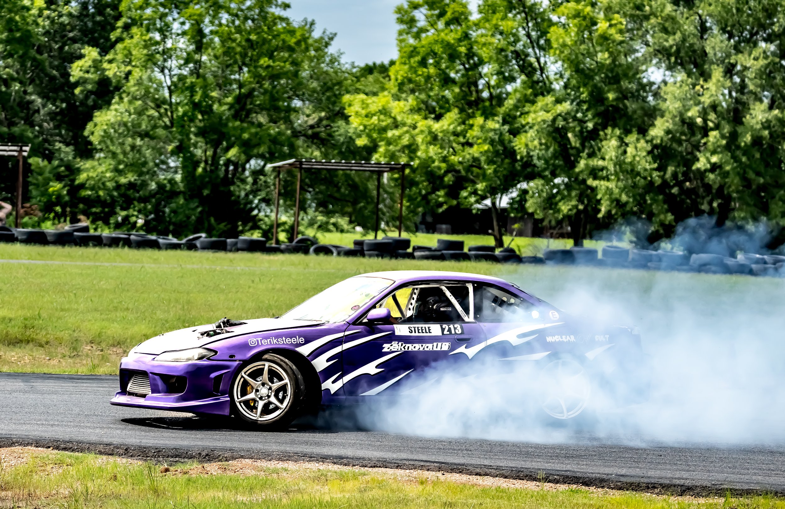 A purple race car with a driver inside is drifting around a corner, creating smoke from the tires on a racetrack with tire barriers in the background and green trees.
