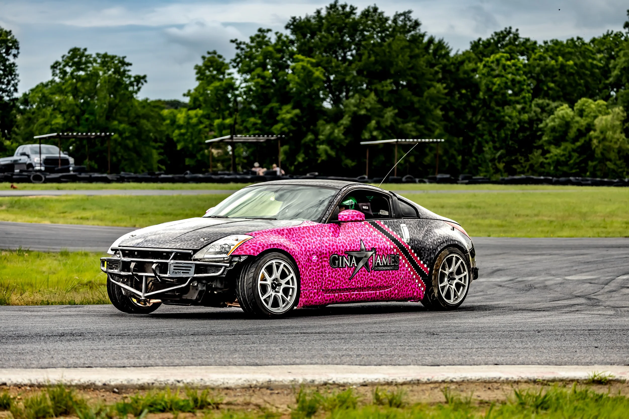 A race car with a pink and black leopard print design, featuring the name 'Gina James Entertainment LLC,' driving on a racetrack with green grass and trees in the background.
