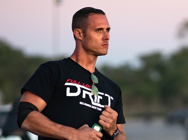 Man with short dark hair wearing a black T-shirt, holding sunglasses and a watch, standing outdoors with a blurred background.