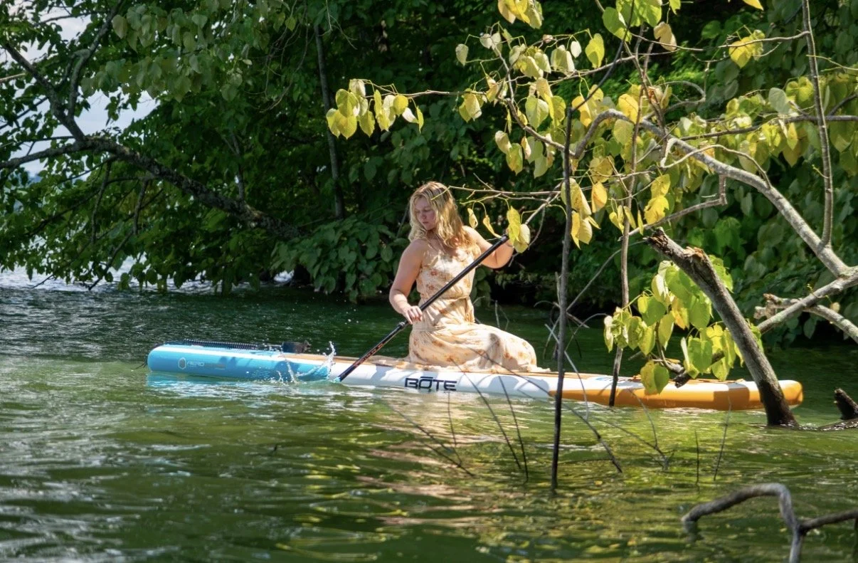 Young woman in a floral dress paddleboarding on a calm river surrounded by lush green trees with yellow leaves. Charlotte NC SUP Rentals